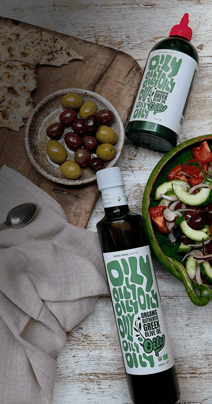 Two bottles of Oily next to a spread of hummus and greek salad