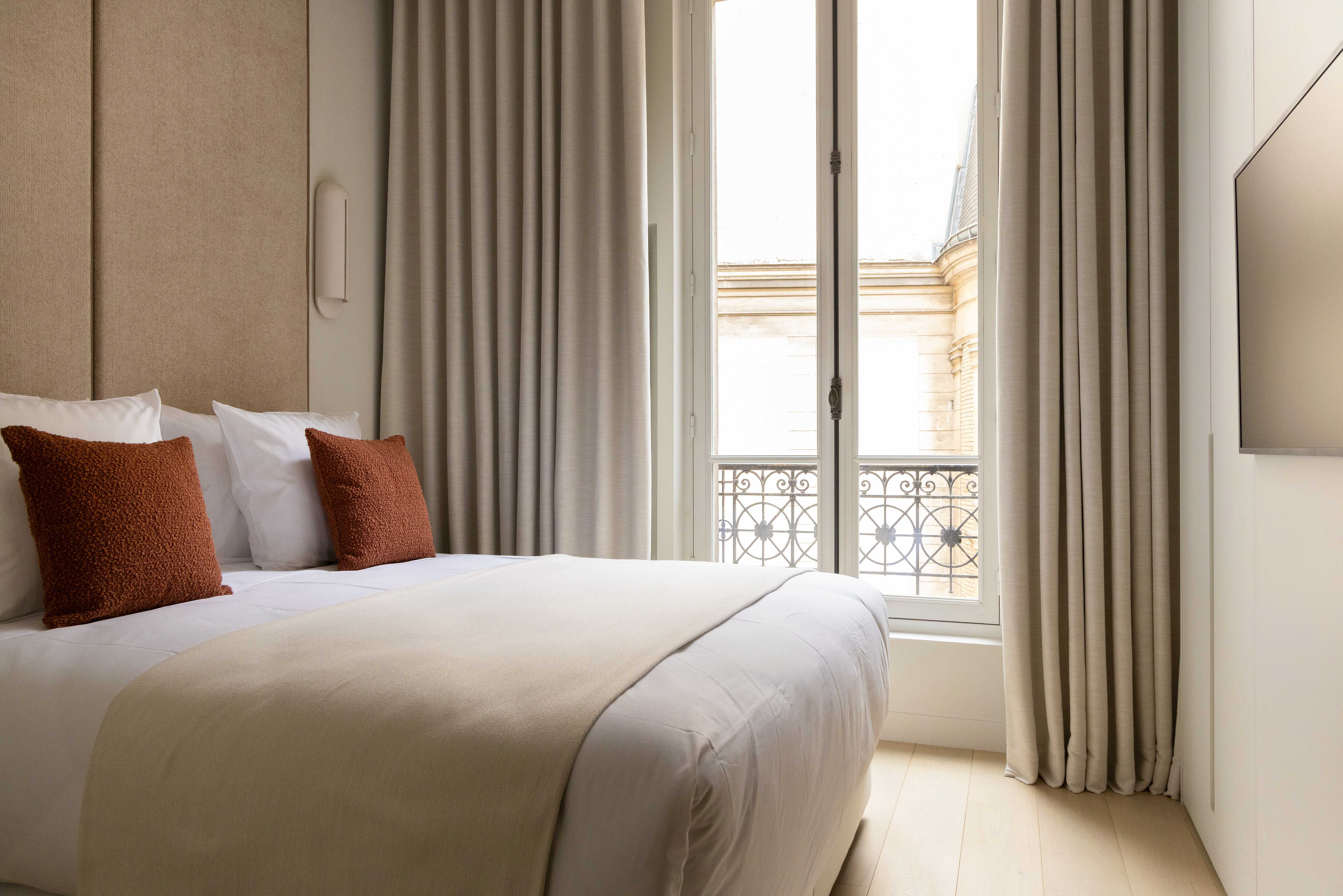 Chambre lumineuse avec lit double, coussins terracotta, tete de lit sable, fenetre ouvrant sur vue parisienne