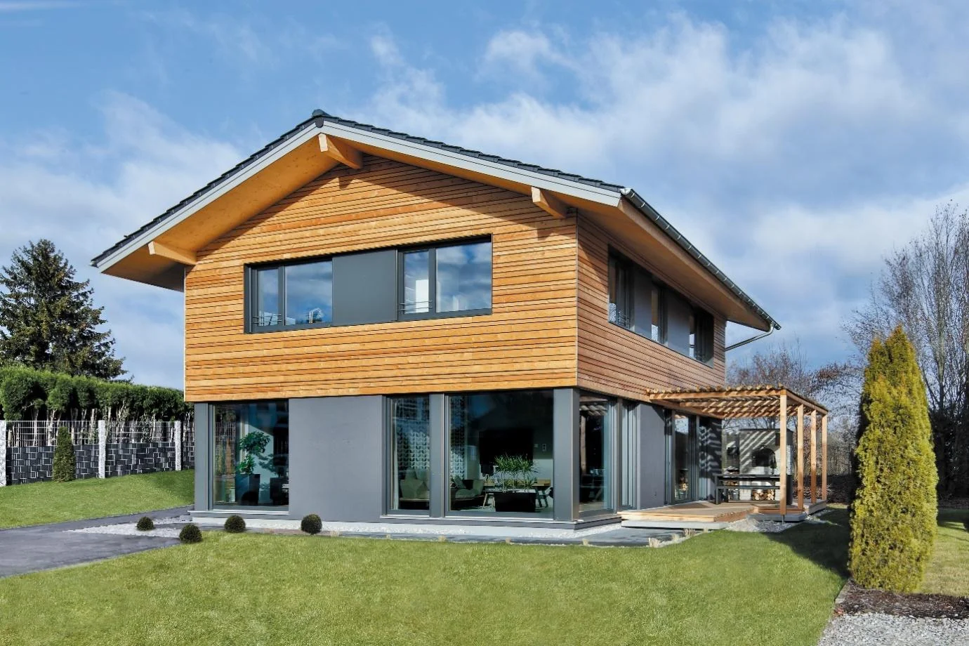 Modernes Holzhaus mit großen Fenstern, Dachüberstand, Holzpergola und gepflegtem Garten.