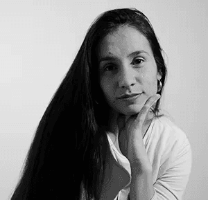 Professional headshot of a brunette woman in a white shirt, arms crossed, on a clean minimal background.