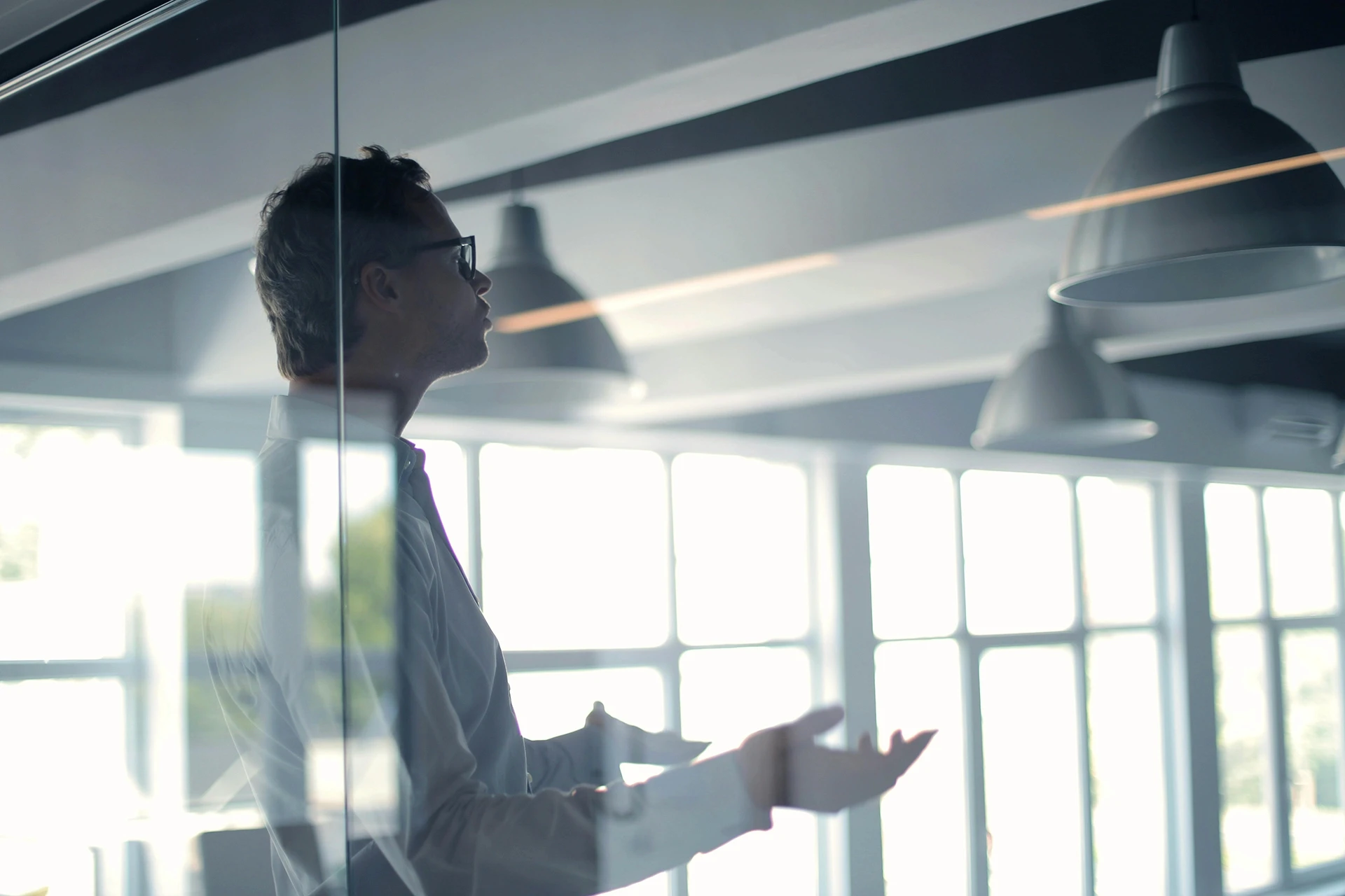 Business man in white shirt and glasses speaking in glass office with large low hanging lampshades