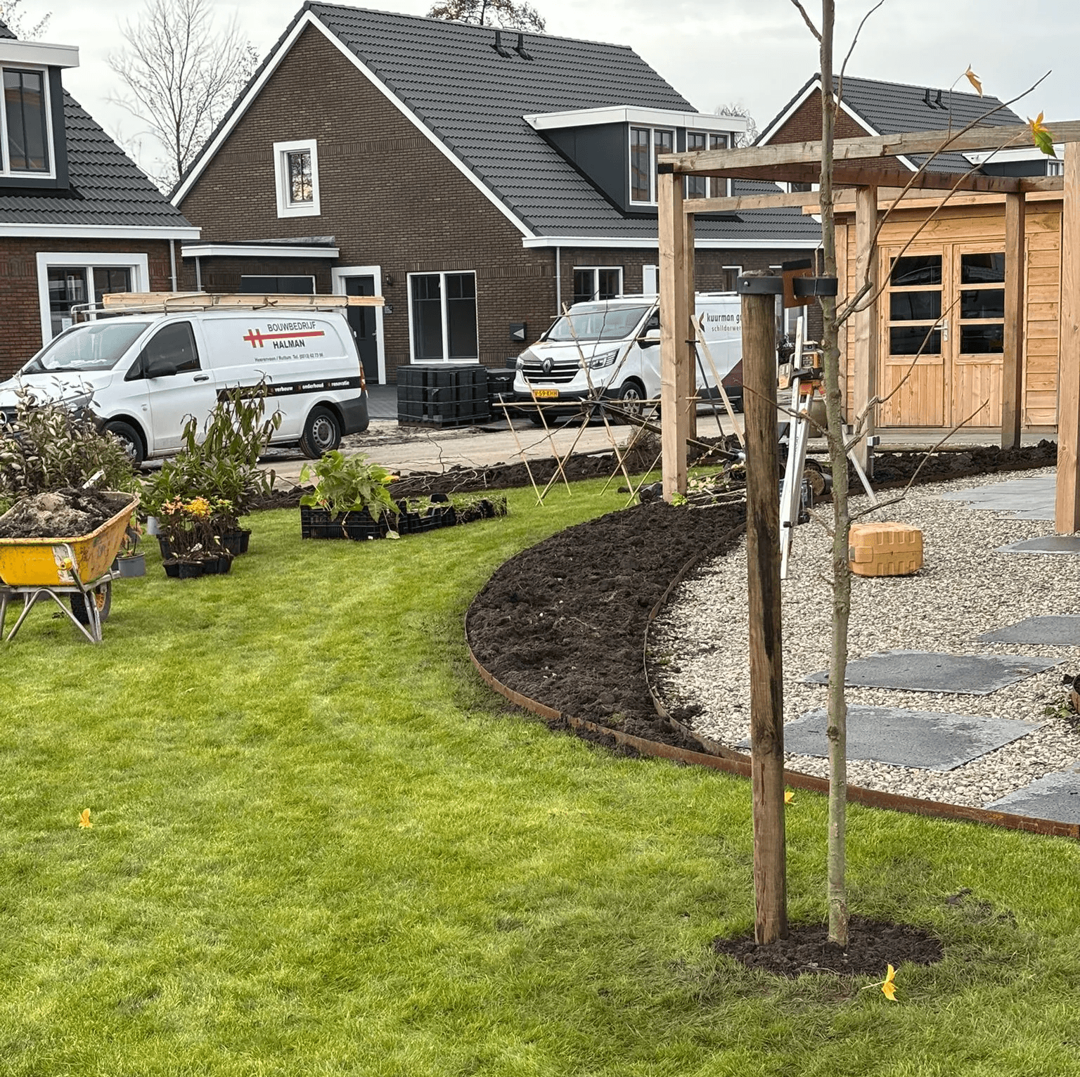 Nieuw aangelegd gazon met organische tuinrand van aarde en grind, jonge boom met steunpaal en houten tuinhuis in aanlegfase.