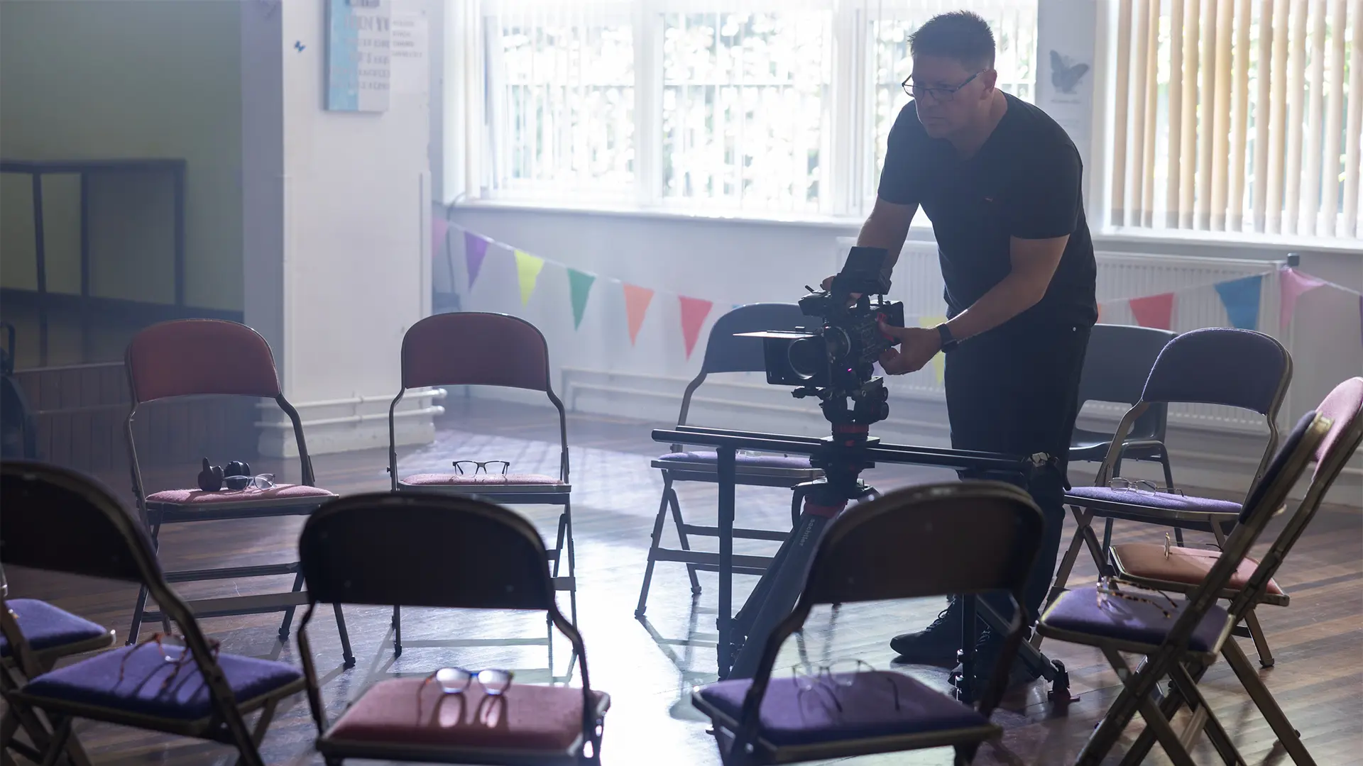 Camera operator setting up a shot in a community hall with chairs arranged in a circle.