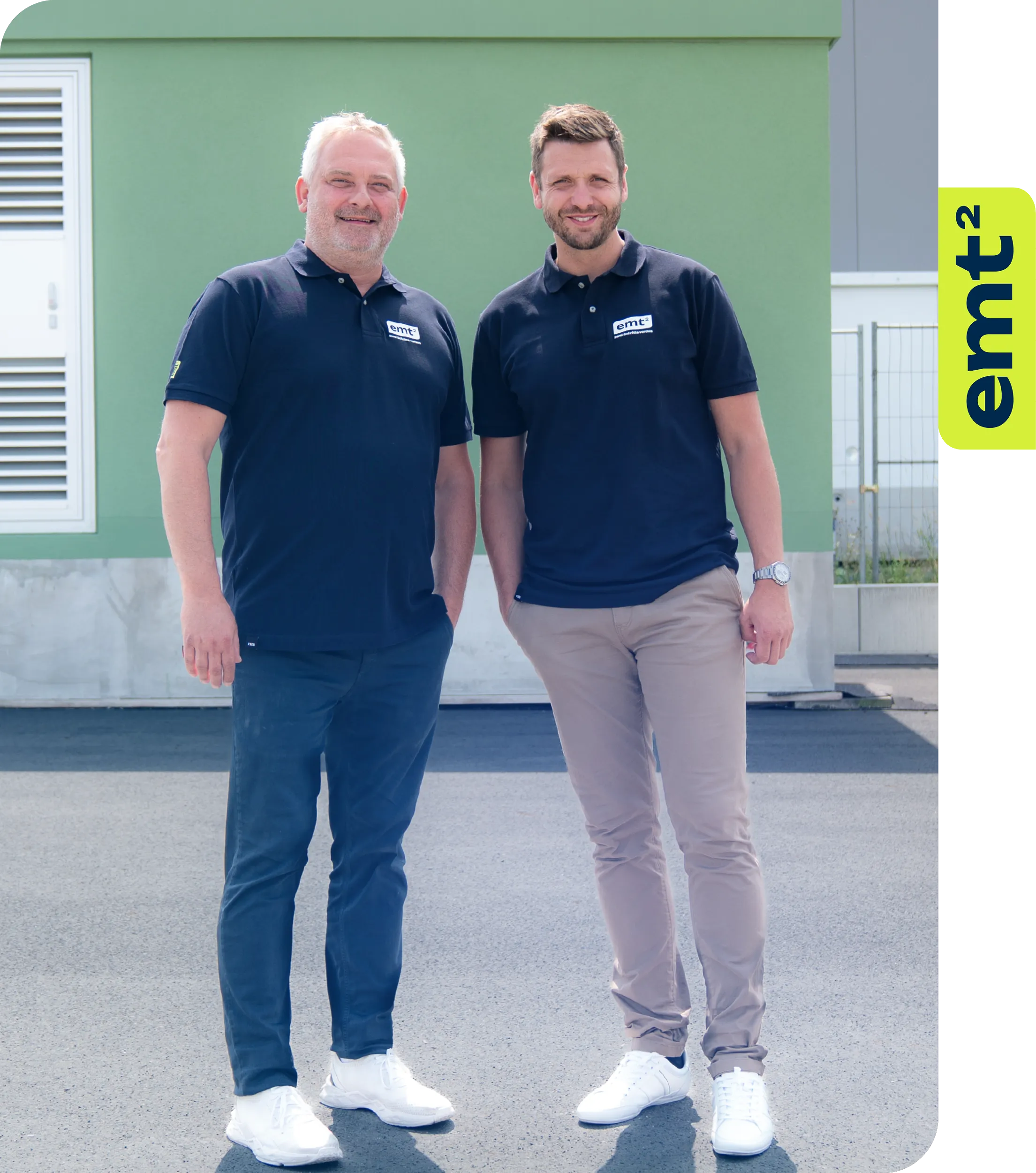 Zwei Männer in Poloshirts und Hosen vor einem Gebäude mit dem Logo „emt²“.