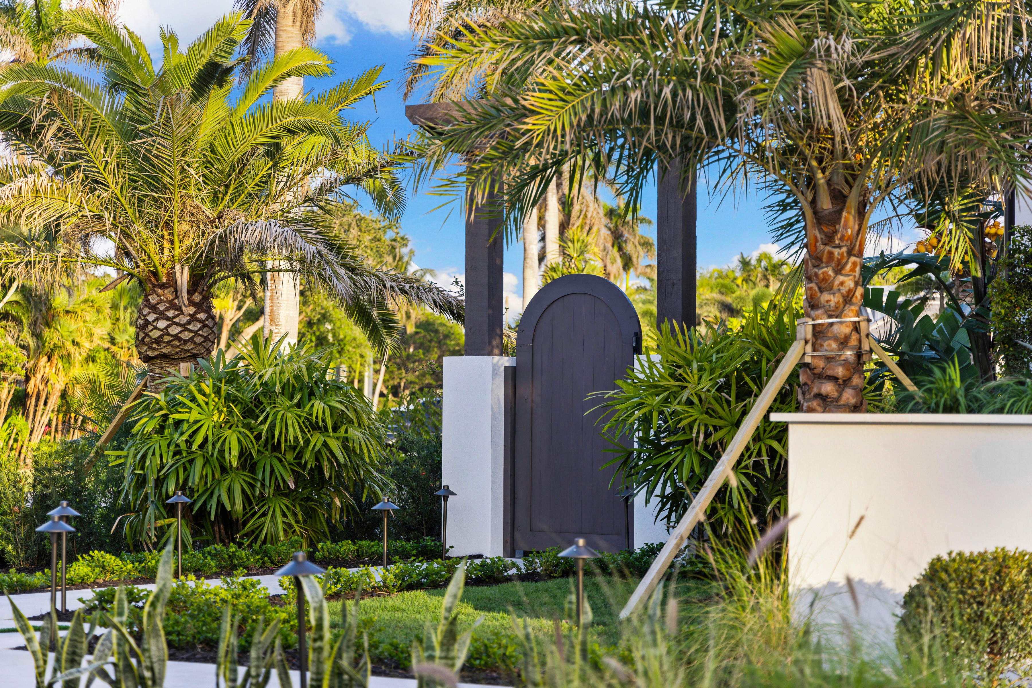 a dark wood gate with white pillars in a nature-like setting with palm trees, bushes, and other landscaping