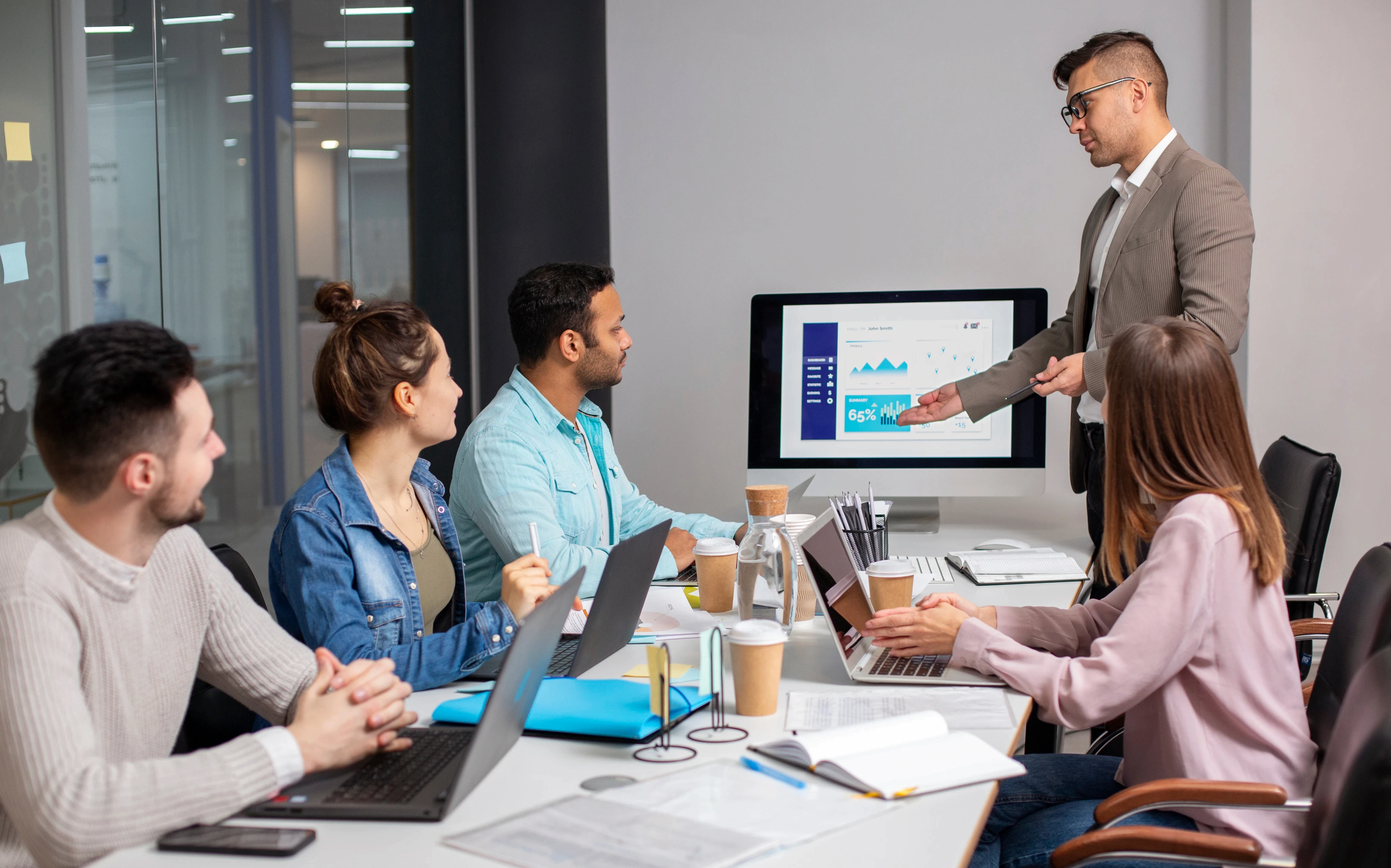 A diverse group of young business professionals in a modern office, listening to a presentation of data analytics on a computer monitor and taking notes.