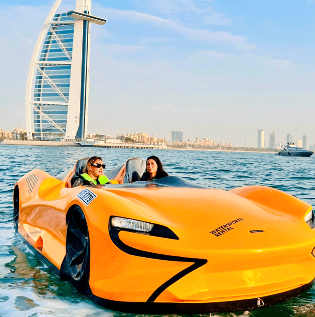 Two women riding a jet car with Dubai's iconic sail-shaped building in the background.