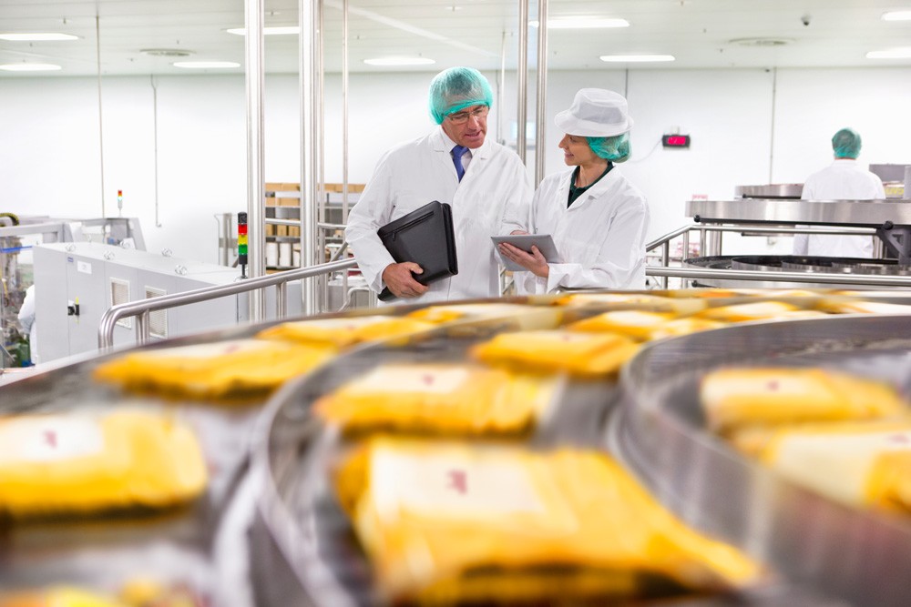 Two food processing experts in white lab coats and hairnets discuss quality control as egg products move along a conveyor.