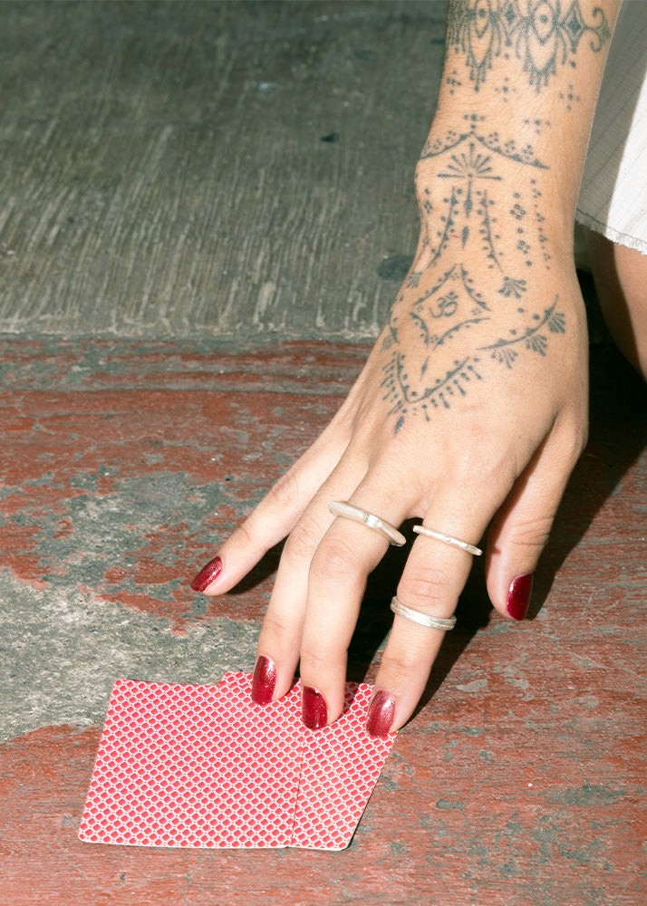 Hand with tattoos and red nail polish holding a deck of cards wearing silver jewelry