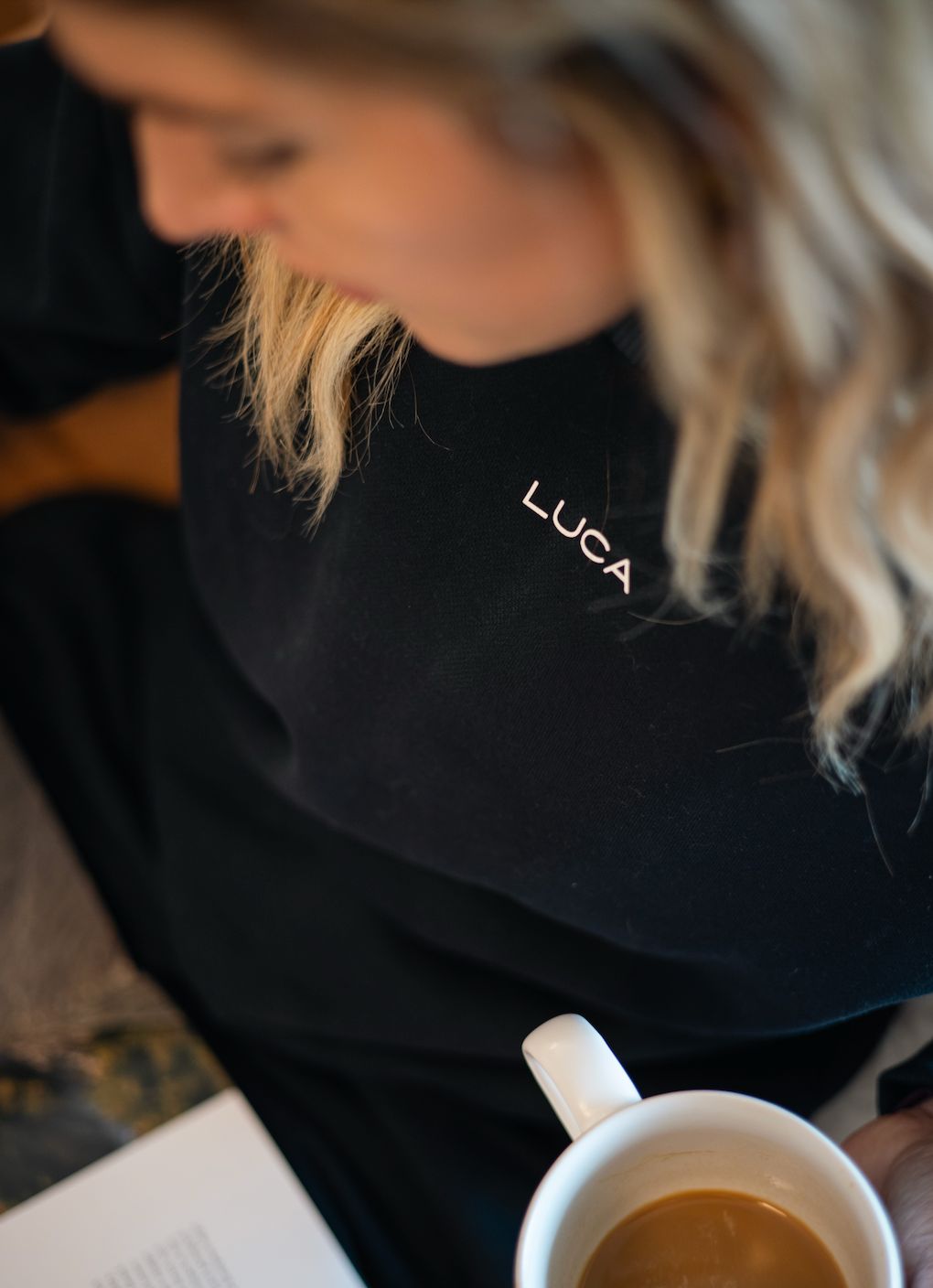 A-women-wearing-a-black-sweatshirt- with-a-small-“LUCA”-logo-looks-down- while-holding-a-cup-of-coffee- creating-a-cozy-relaxed-moment