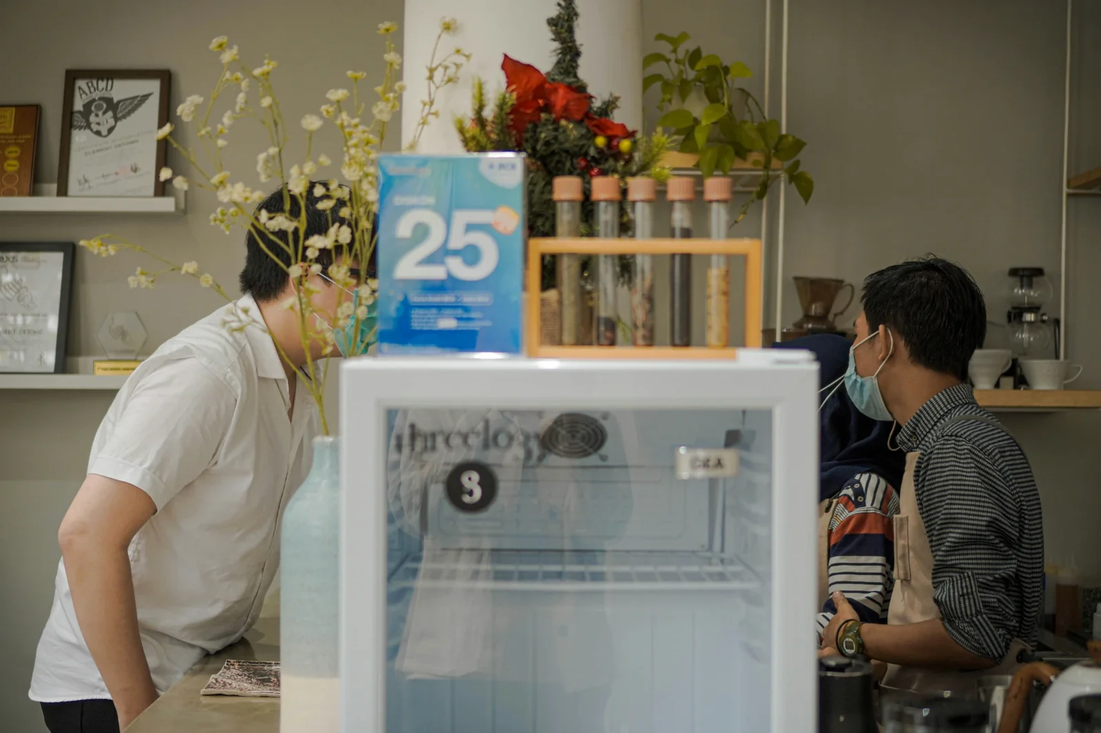 Two people interact at a refrigerated display case marked "3" in a bright retail space decorated with plants and flowers.