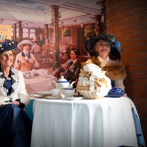 Two women in vintage attire sit at a table with teapots and decorative items, backed by a mural of a historic dining scene.