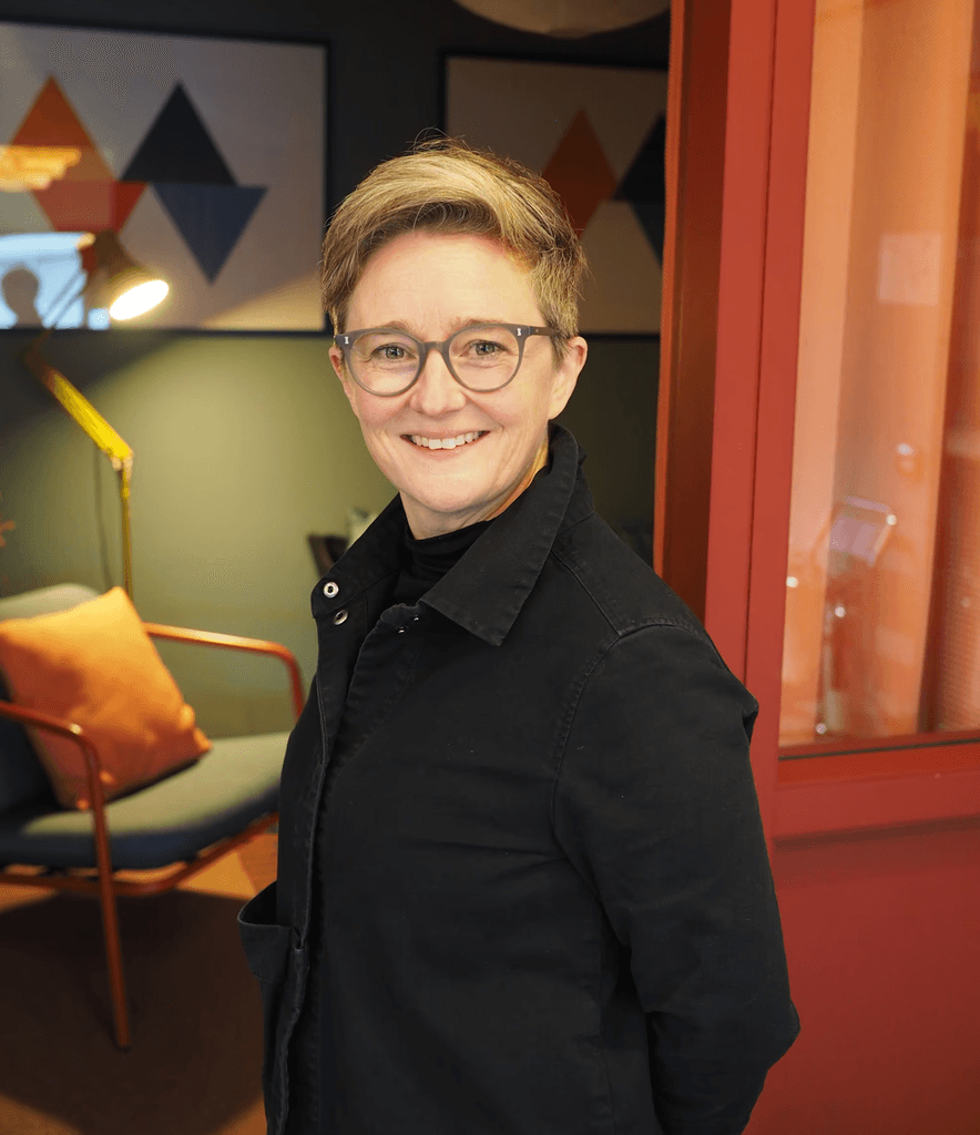 A person stands indoors in a warmly lit room, wearing a dark jacket and facing the camera. Behind them is a modern seating area with a green upholstered chair, a floor lamp, patterned wall art, and a red-framed glass partition, creating a cozy, contemporary interior.