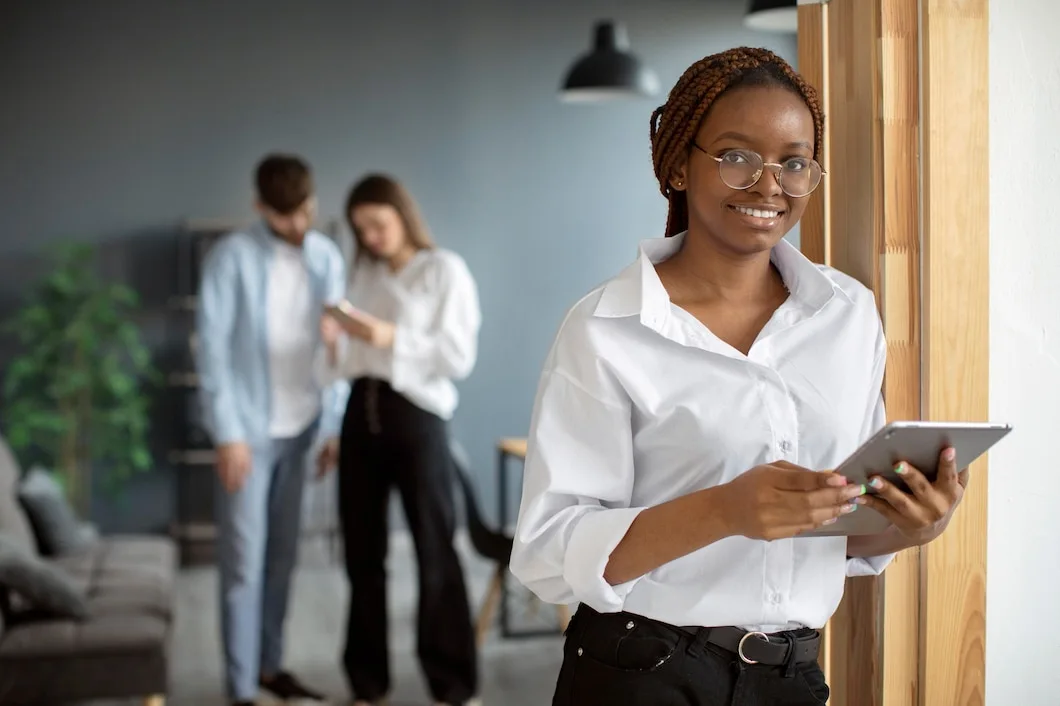 A imagem mostra uma mulher jovem, sorridente, vestindo uma camisa branca e segurando um tablet. Ela usa óculos e está encostada em uma parede de madeira, em um ambiente de escritório moderno. Ao fundo, um casal de colegas de trabalho parece engajado em uma conversa ou atividade, desfocado em segundo plano. A composição sugere um ambiente de trabalho colaborativo e diversificado.