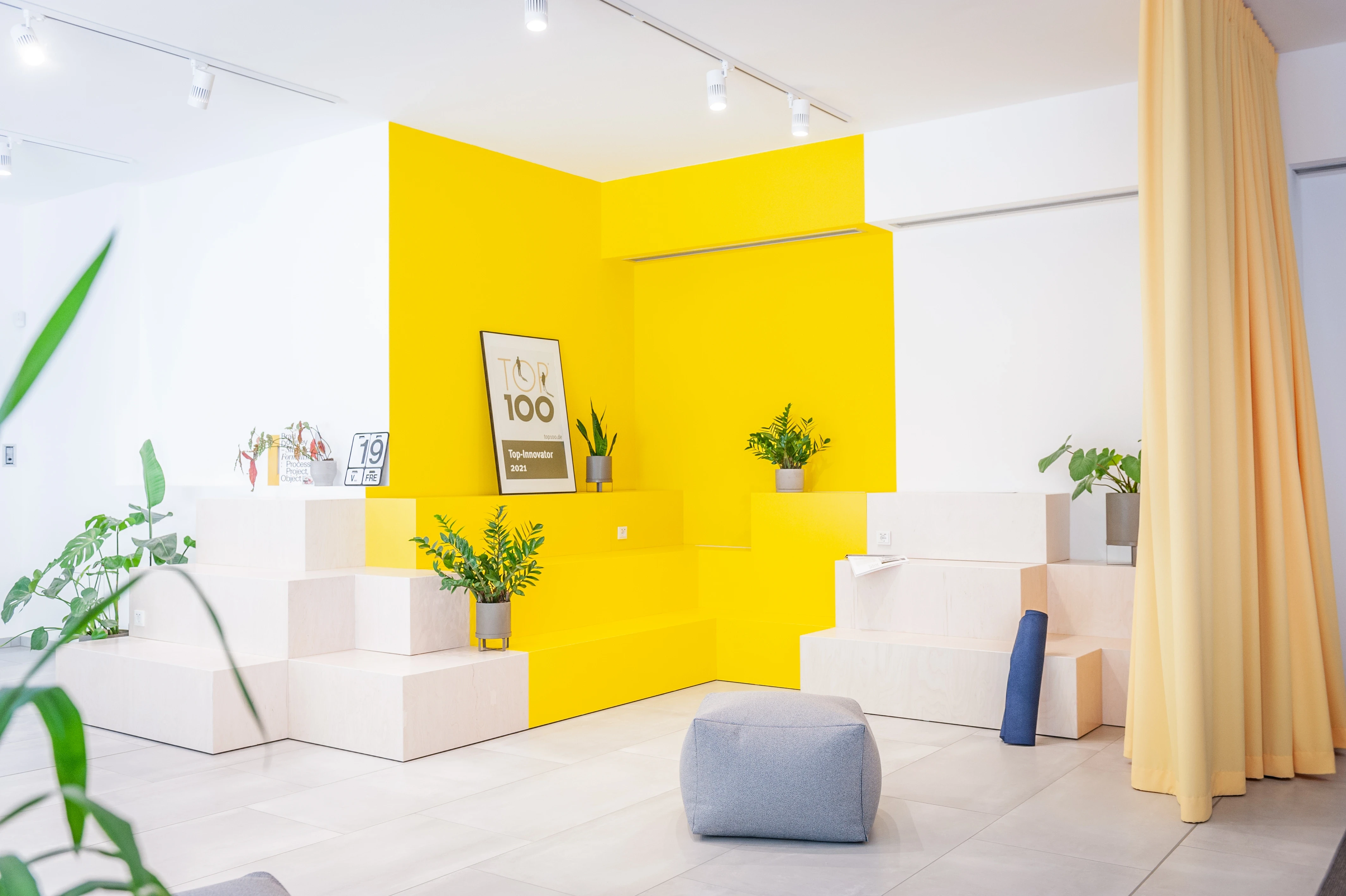Reception area at EHEALTH-TEC with a bright yellow corner wall and matching stepped seating blocks in pale stone, potted plants, a grey pouf, a blue bolster cushion and a yellow curtain.