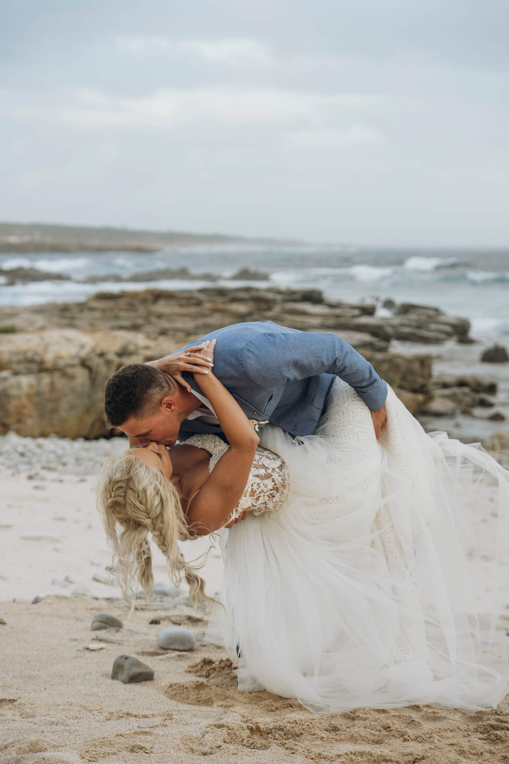 Groom dipping bride while kissing on beach with ocean backdrop at Mostertshoek