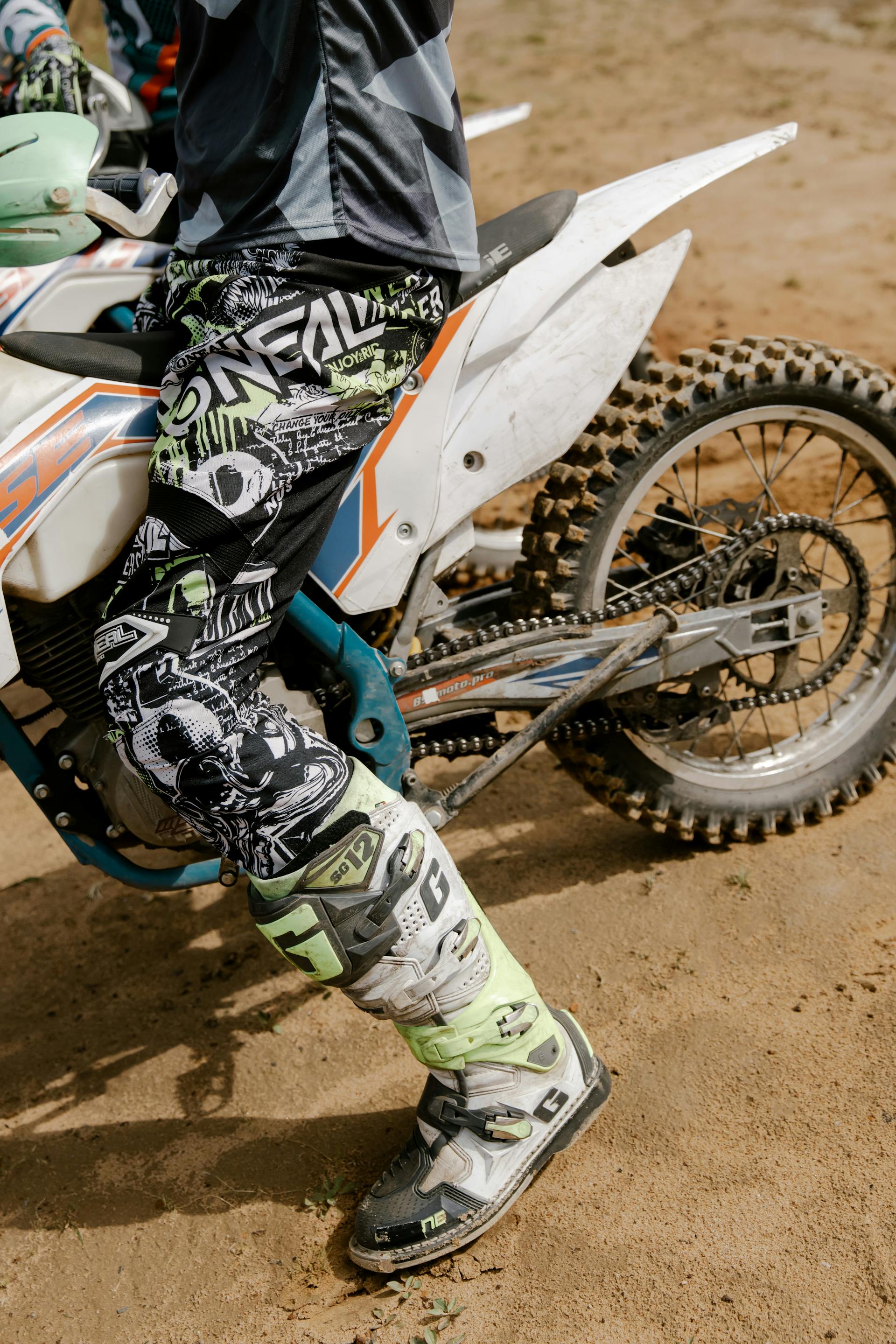 A close-up side view of a rider's leg wearing patterned black and white motocross pants and a sturdy white and neon yellow riding boot while seated on a white dirt bike.