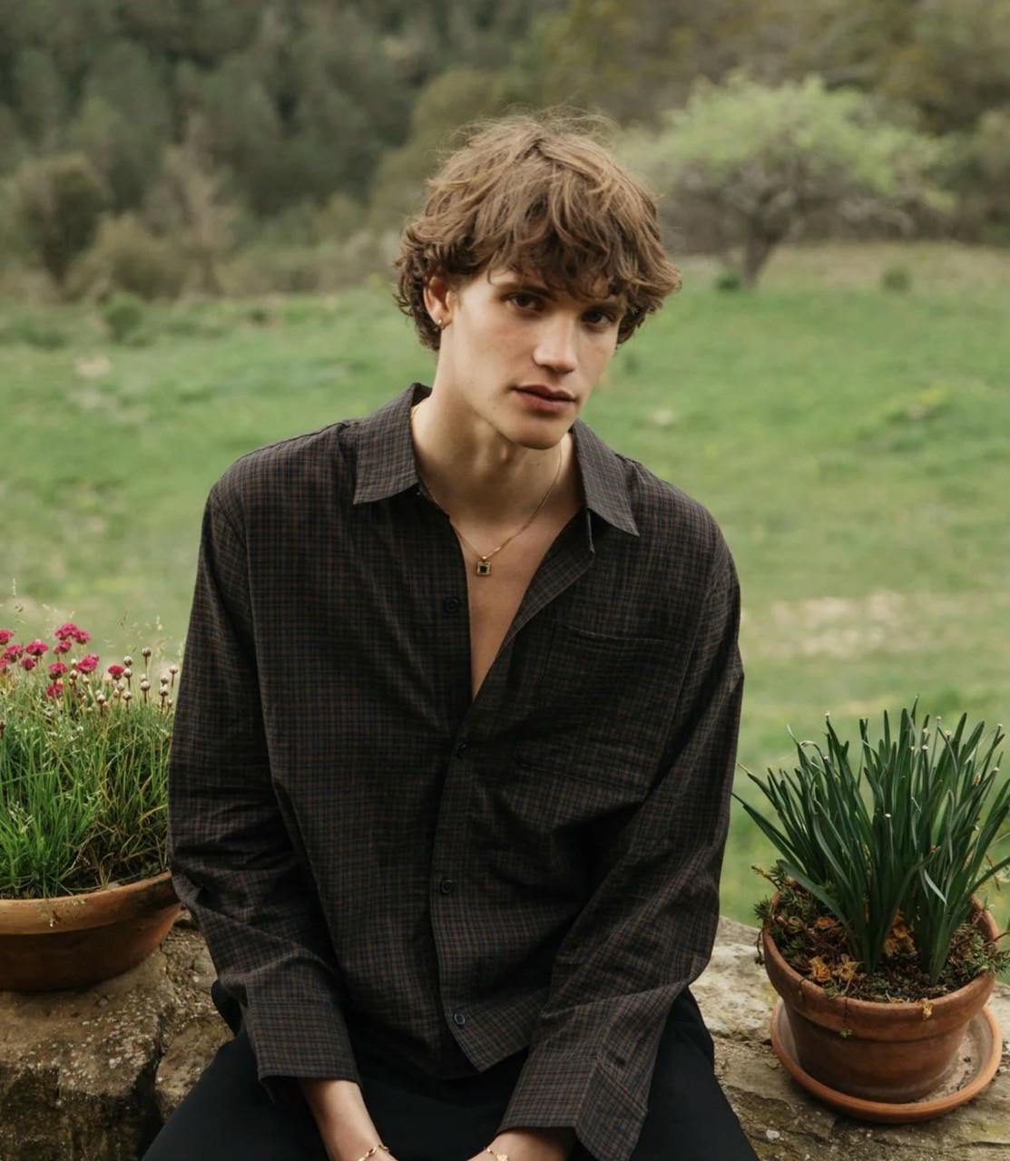 A young man sitting in a garden wearing 33mm jewelry and apparel