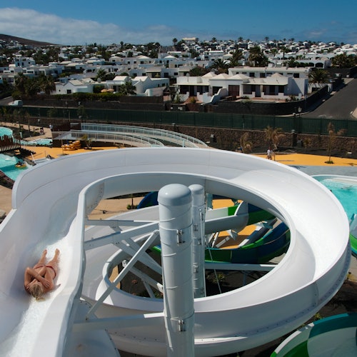 在一个水上乐园中，一位游客从白色的扭曲滑梯滑下，背景是一个住宅区。
