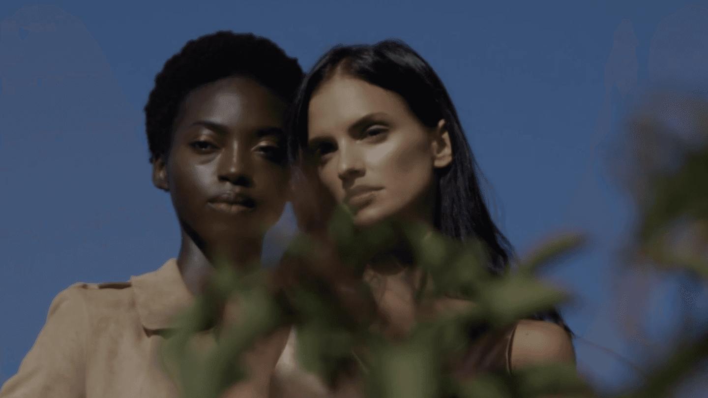 Two women standing closely together outdoors under a clear blue sky, looking directly at the camera, with blurred green leaves partially covering the foreground.