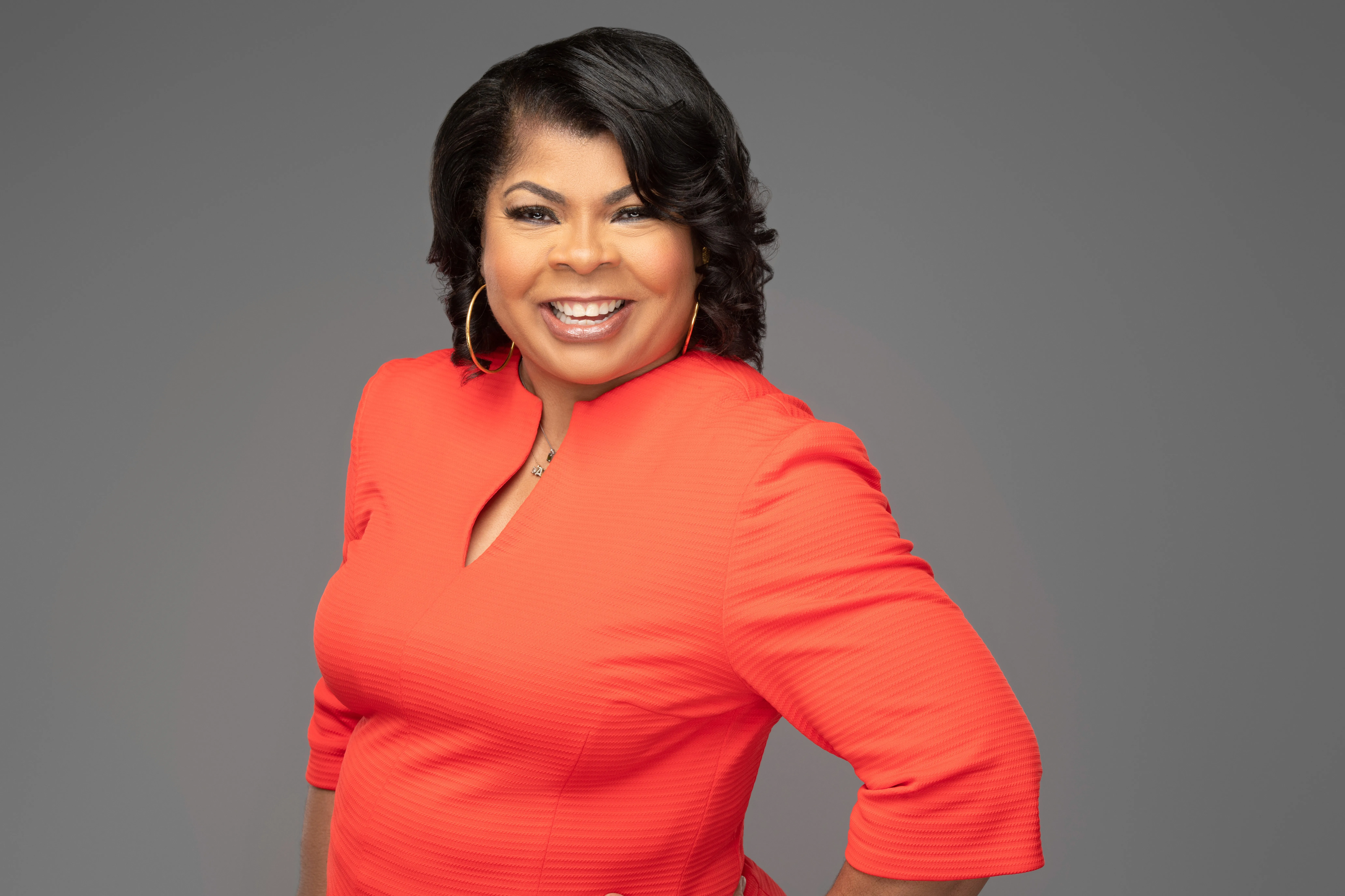 April Ryan with medium-length dark hair smiles confidently, wearing a bright orange dress with three-quarter sleeves. She stands against a plain gray background, posing with one hand on her hip.