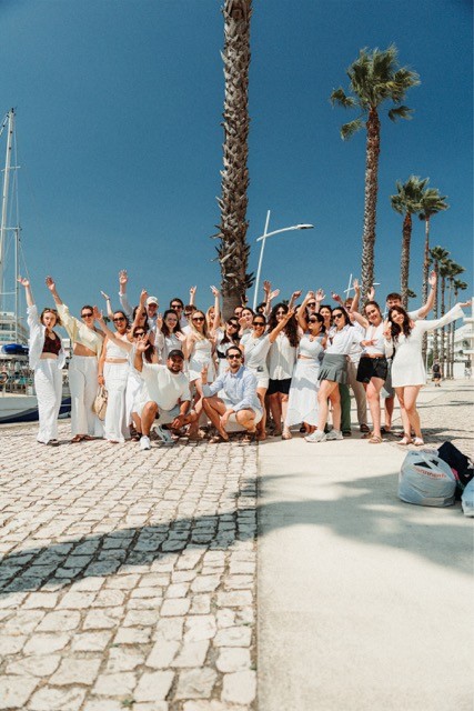 A group of eight people stands together on a walkway by the beach at sunset, smiling and posing for the camera.