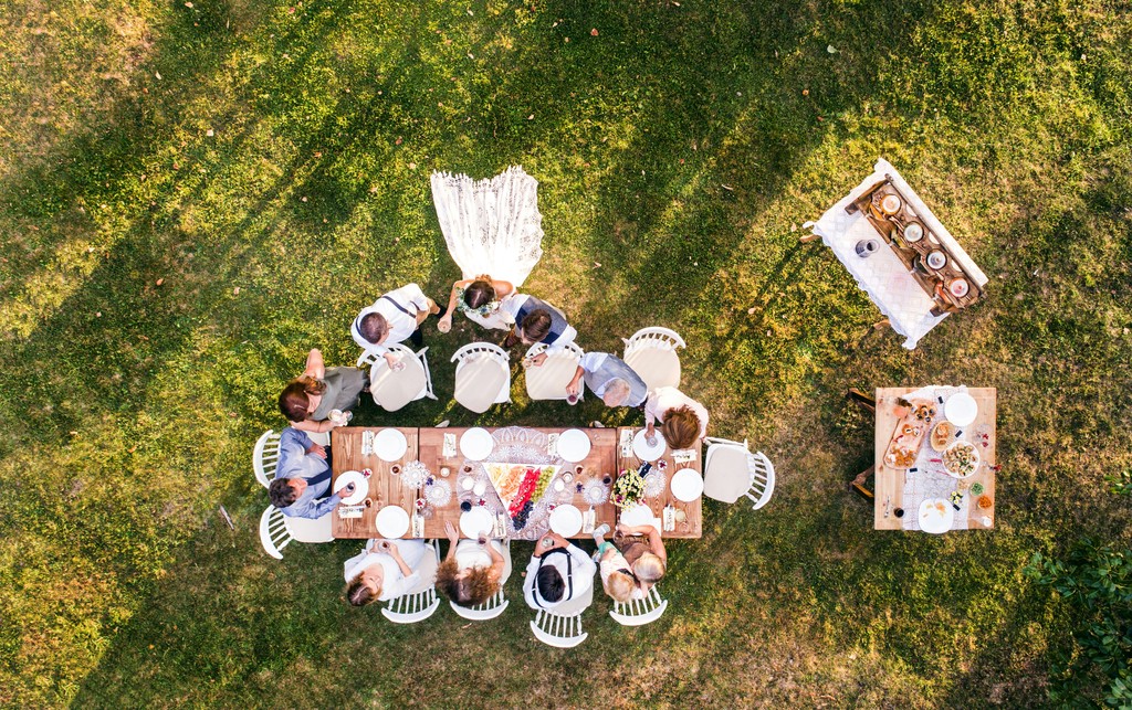 An aerial view of a wedding reception in a lush green garden, where guests are seated around a long, decorated table enjoying an outdoor meal, with additional food tables set up nearby.