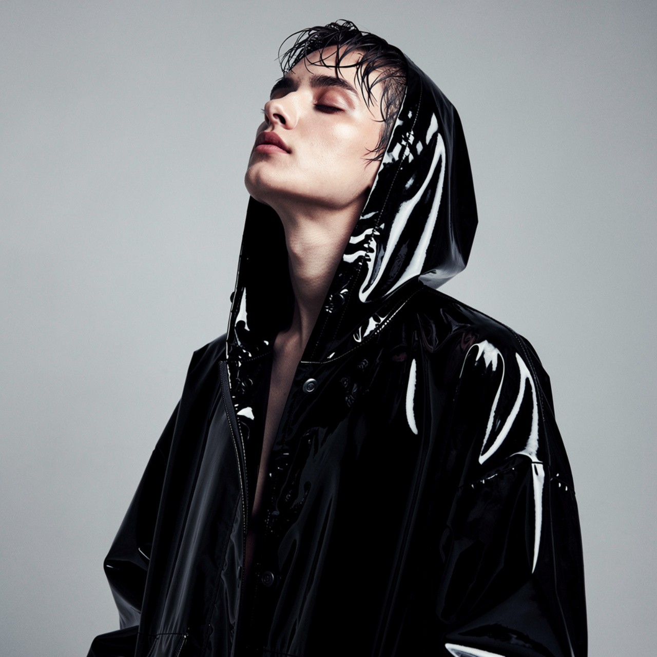 Person in a shiny black hooded jacket, tilting head back, eyes closed; conveys a mood of confidence and contemplation against a gray background.