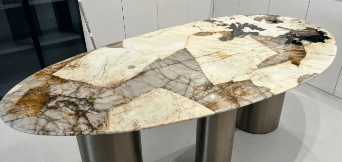 An oval-shaped stone dining table with a complex, patchwork design of different marble slabs in shades of cream, brown, and gray. The table stands on two metallic cylindrical bases.