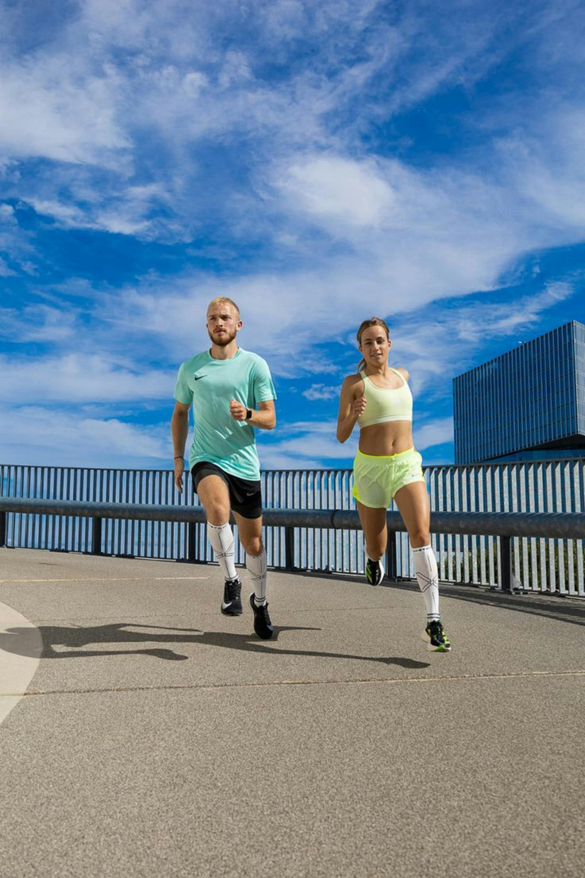 A man and woman in athletic gear jogging side-by-side on a sunny urban bridge.