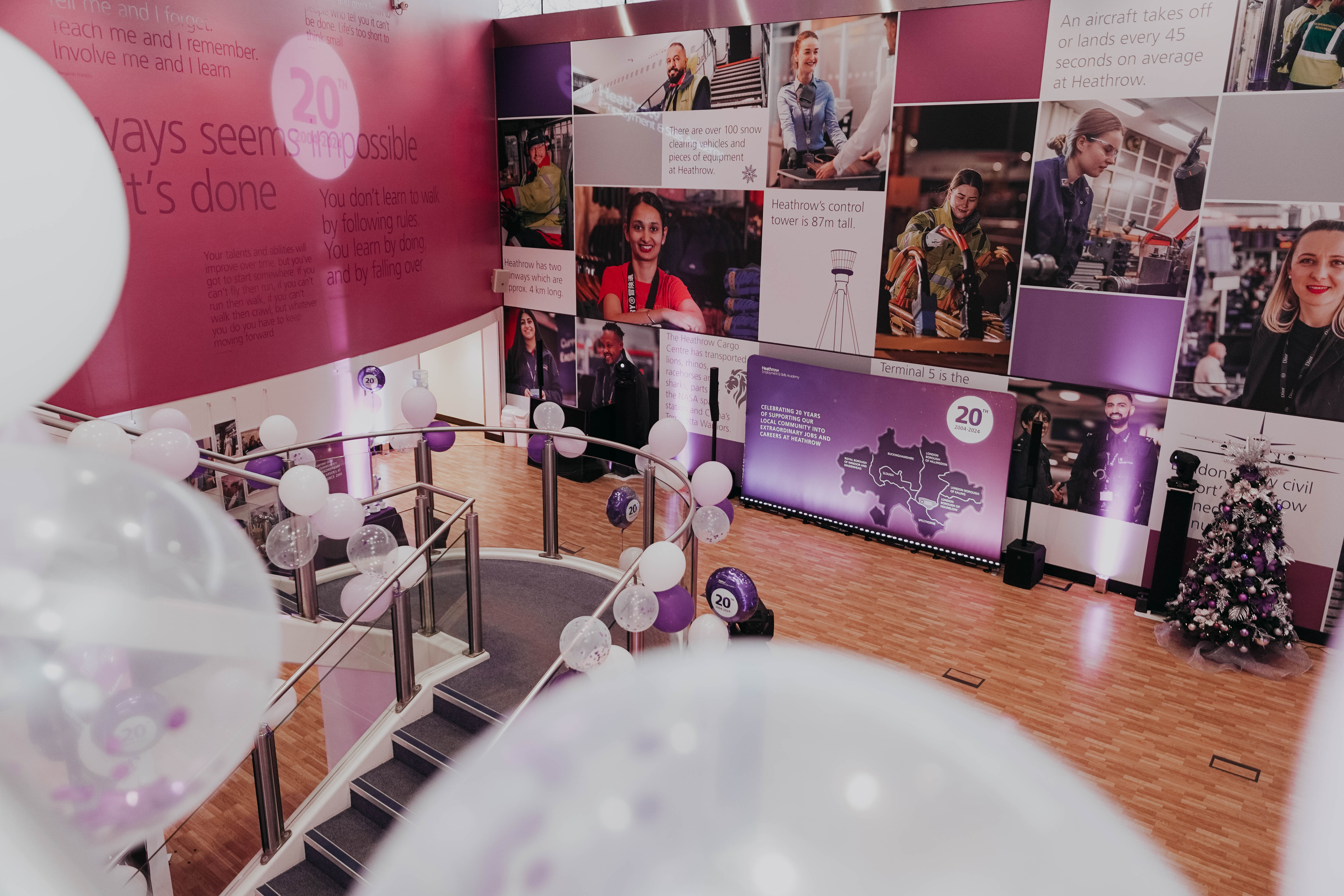 A modern event space decorated with purple and white balloons to celebrate 20 years of Heathrow Academy. A large photo mural covering the walls features images of airport staff and operations, alongside inspirational quotes and a "20 years" logo.