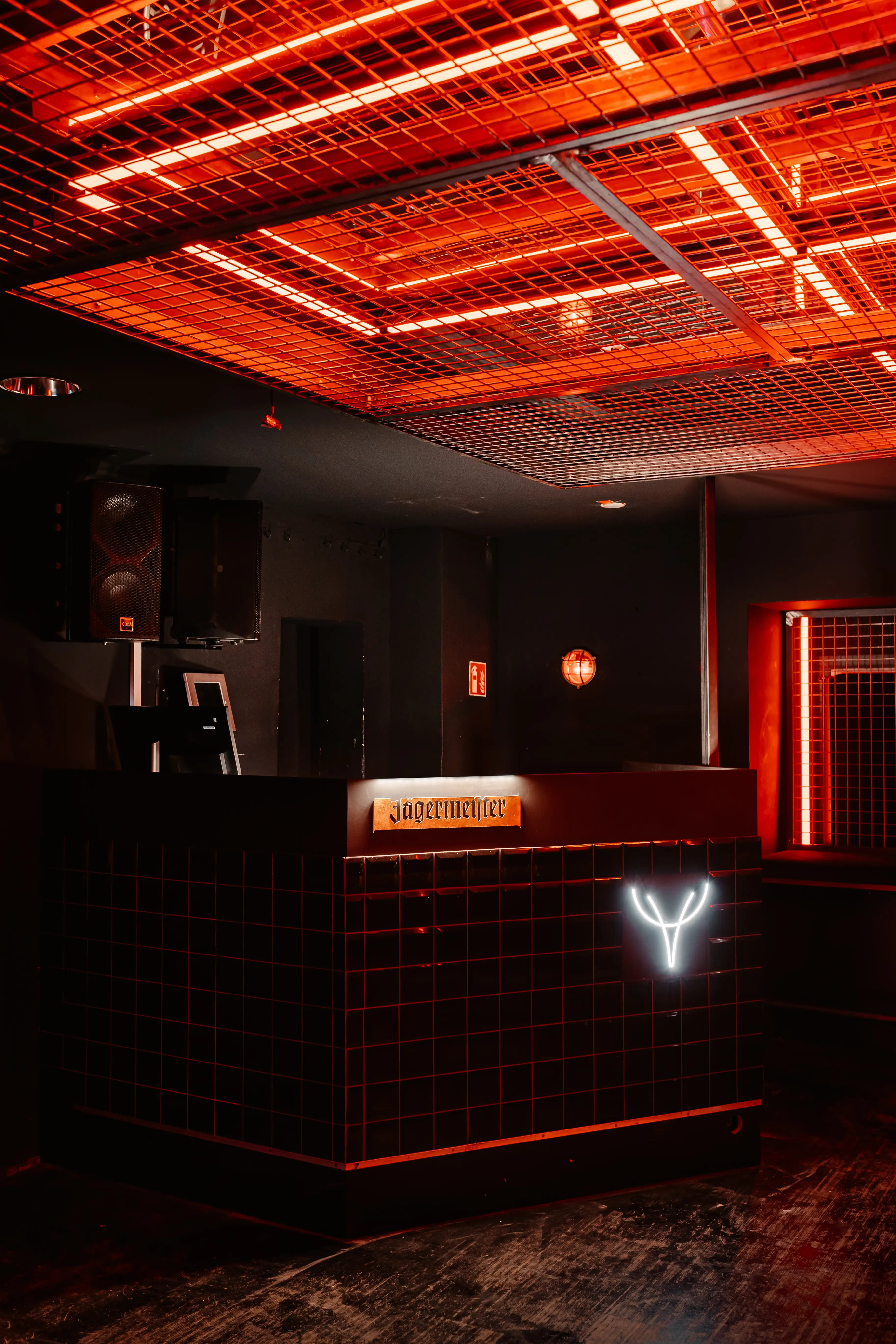 Dark tiled DJ counter with a white neon stag emblem and copper Jägermeister sign at JÄGERMEISTER x CALL ME DRELLA, under a red-glowing grid ceiling with speakers.
