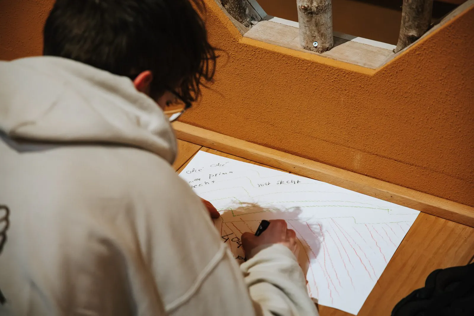 Een man in een lichte capuchontrui schrijft met een stift op een groot vel papier op een houten vensterbank