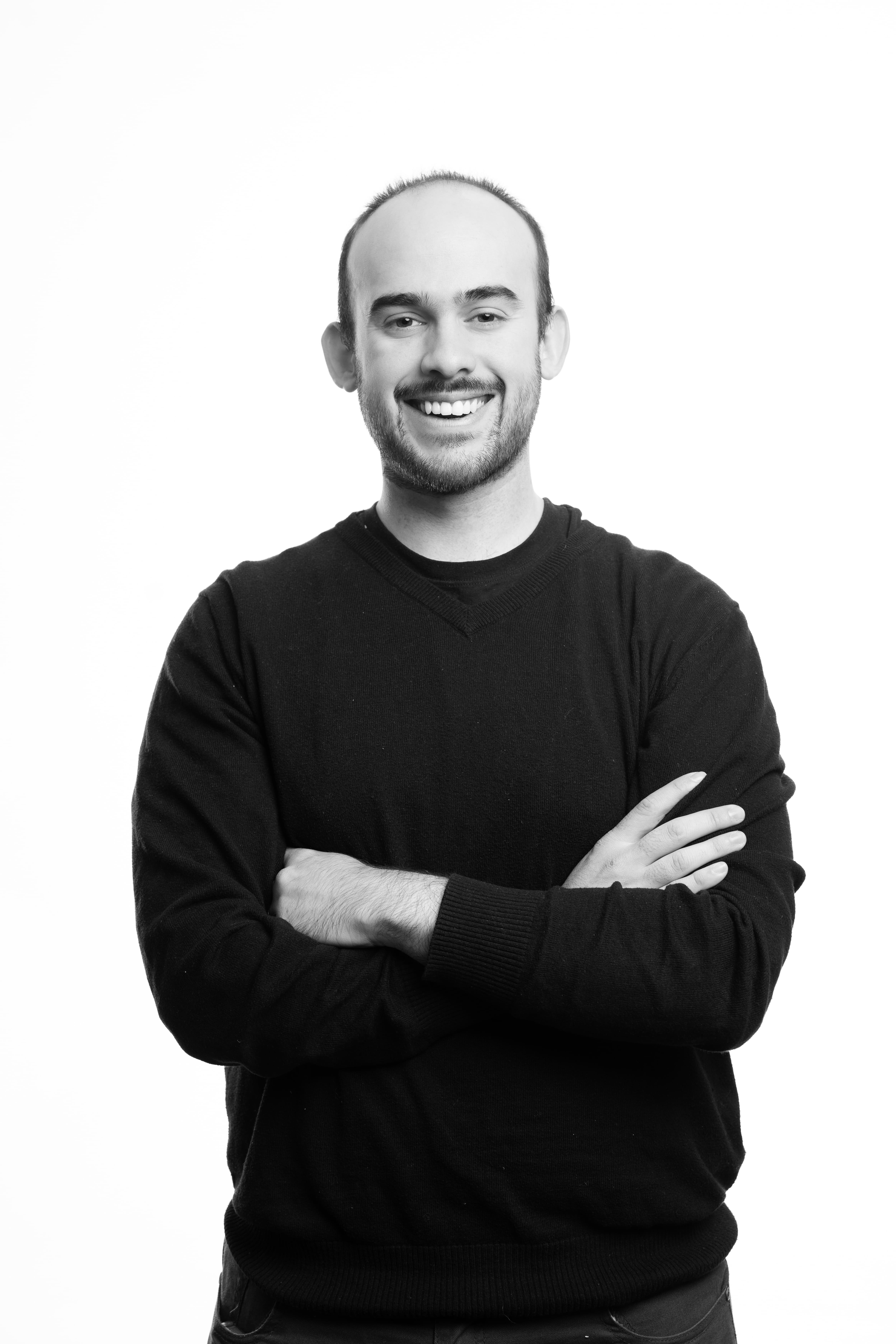 A man with a friendly expression stands confidently with arms crossed. He wears a black sweater against a plain white background, conveying warmth.