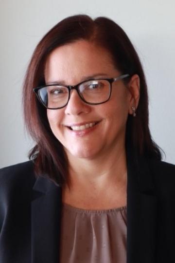 Smiling woman with glasses wearing a dark blazer. She has short, dark hair and a light-colored blouse.