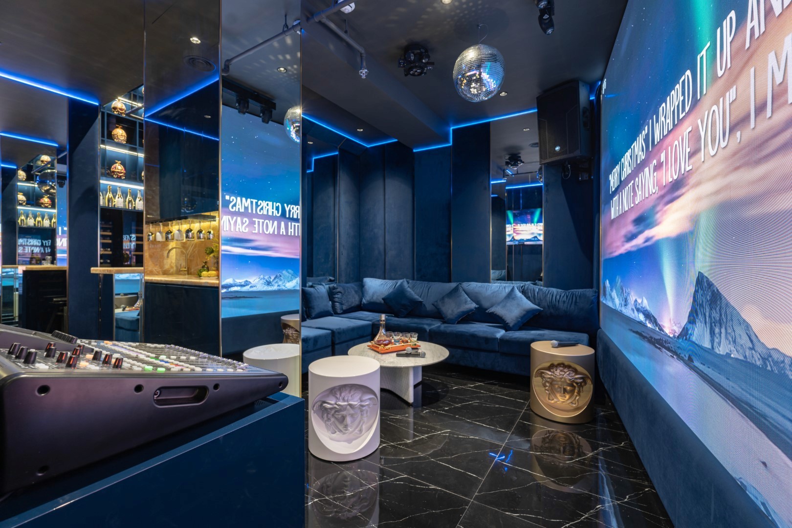 A modern karaoke room features dark blue walls, a reflective ceiling adorned with a disco ball, plush seating, a large illuminated screen displaying song lyrics, and a chic sound mixer on the side, creating an inviting atmosphere for entertainment.