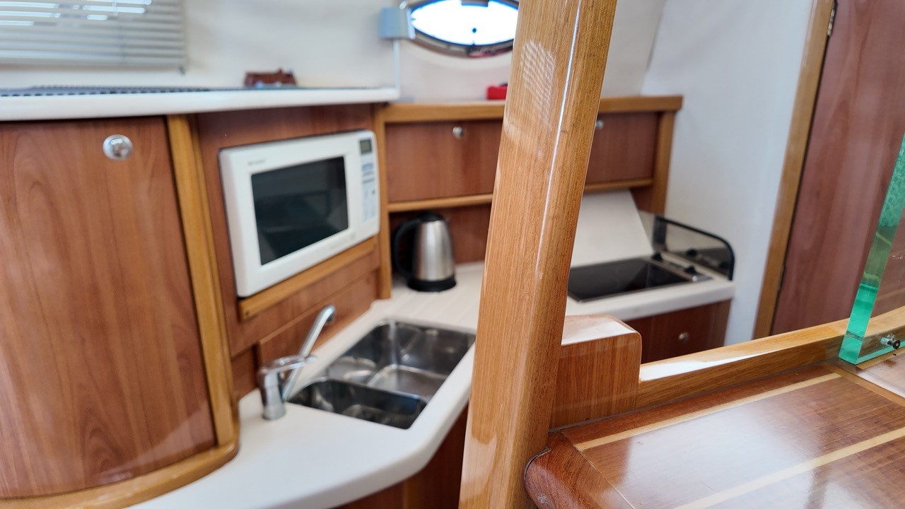 Sealine F37 galley kitchen interior — teak cabinetry, white microwave oven, stainless steel double sink, electric kettle, and two-burner hob with fiddle rails, lit by overhead hatch on a Mediterranean liveaboard motor yacht