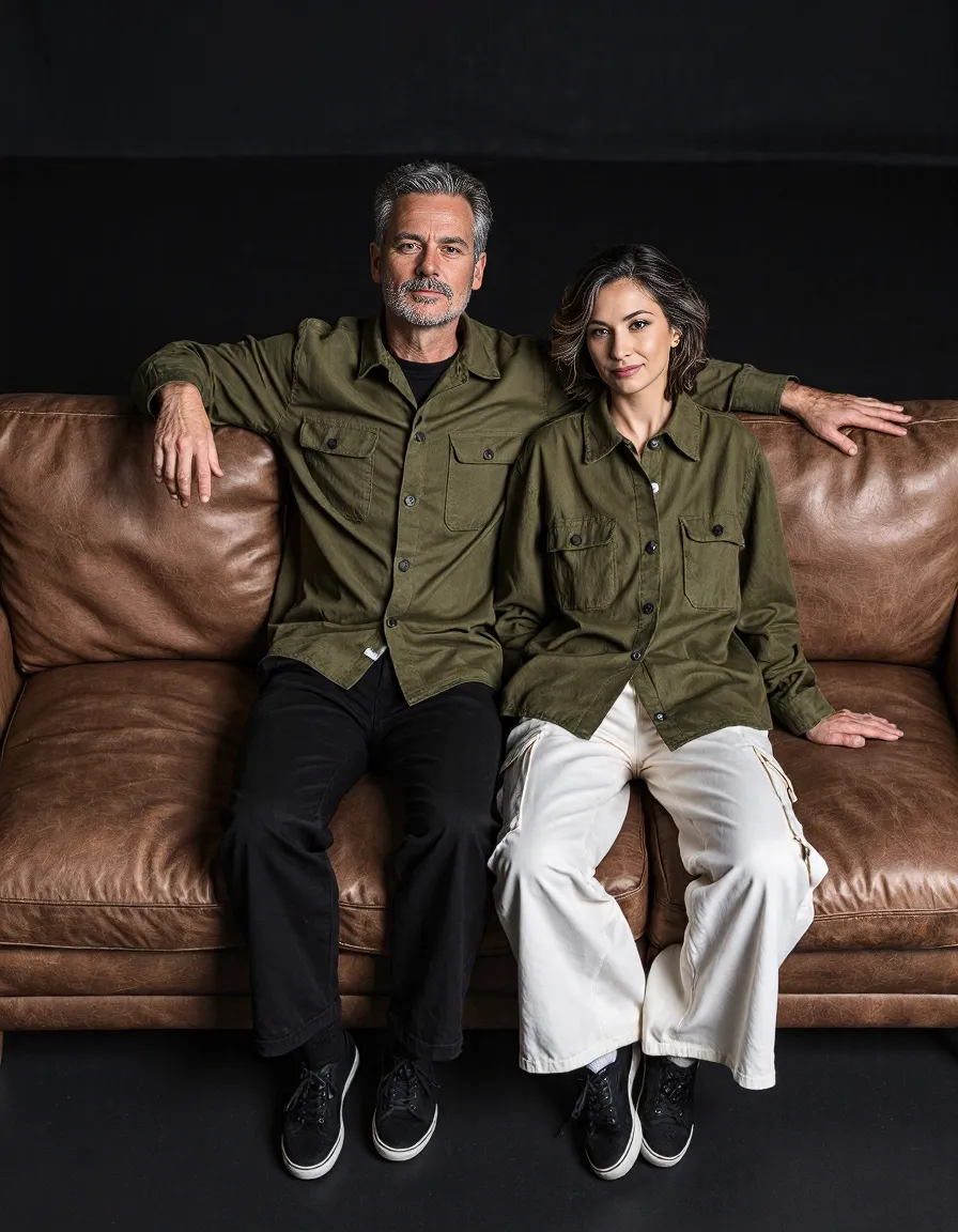 Two people in matching olive green button-up shirts seated on a brown leather couch against a dark backdrop, creating a stylish portrait.
