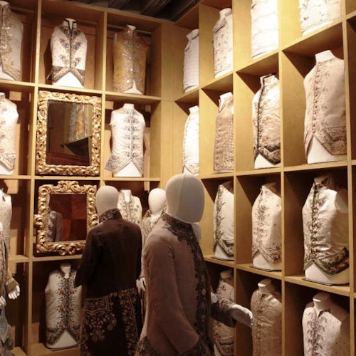 木制展示格中陈列着华丽的历史风格马甲，旁边的人体模特也穿着类似服饰，墙上还挂着一面装饰镜。