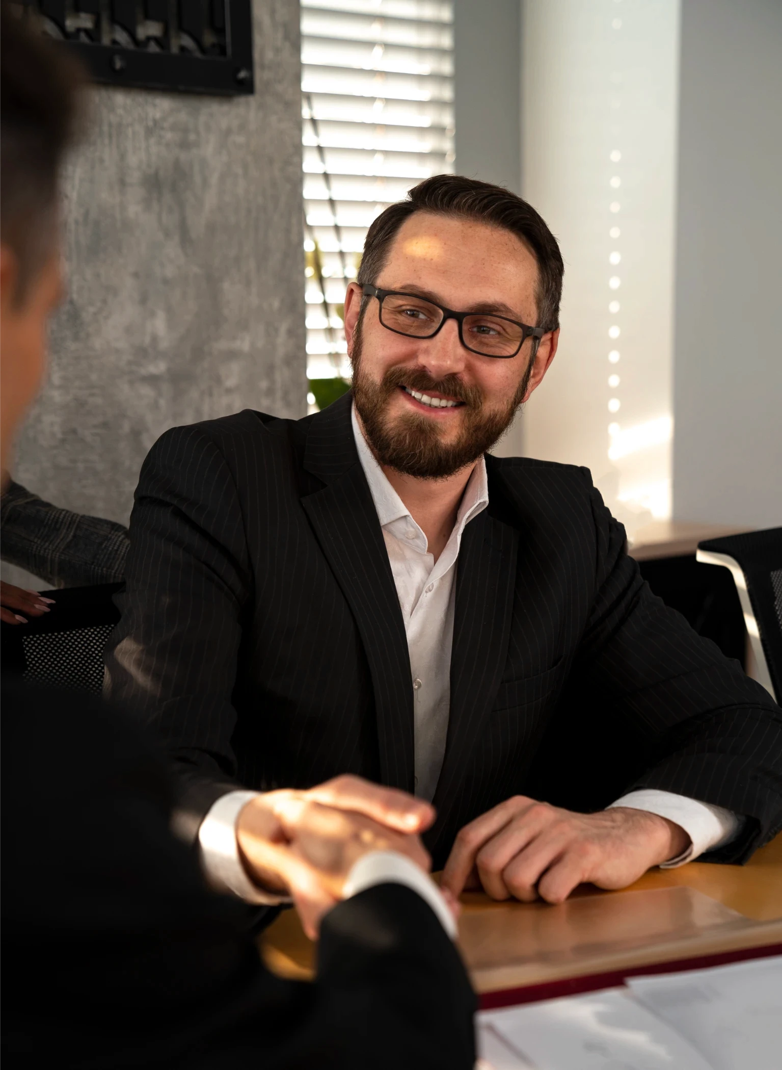 A smiling man in a suit and glasses shakes hands with another person across a table in a sunlit office, conveying a sense of collaboration and agreement.