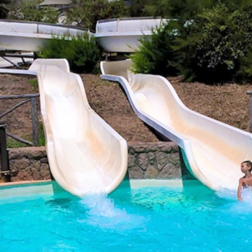 Due scivoli d'acqua si riversano in una piscina turchese, con una persona nell'acqua. Sullo sfondo ci sono cespugli e una recinzione di legno.