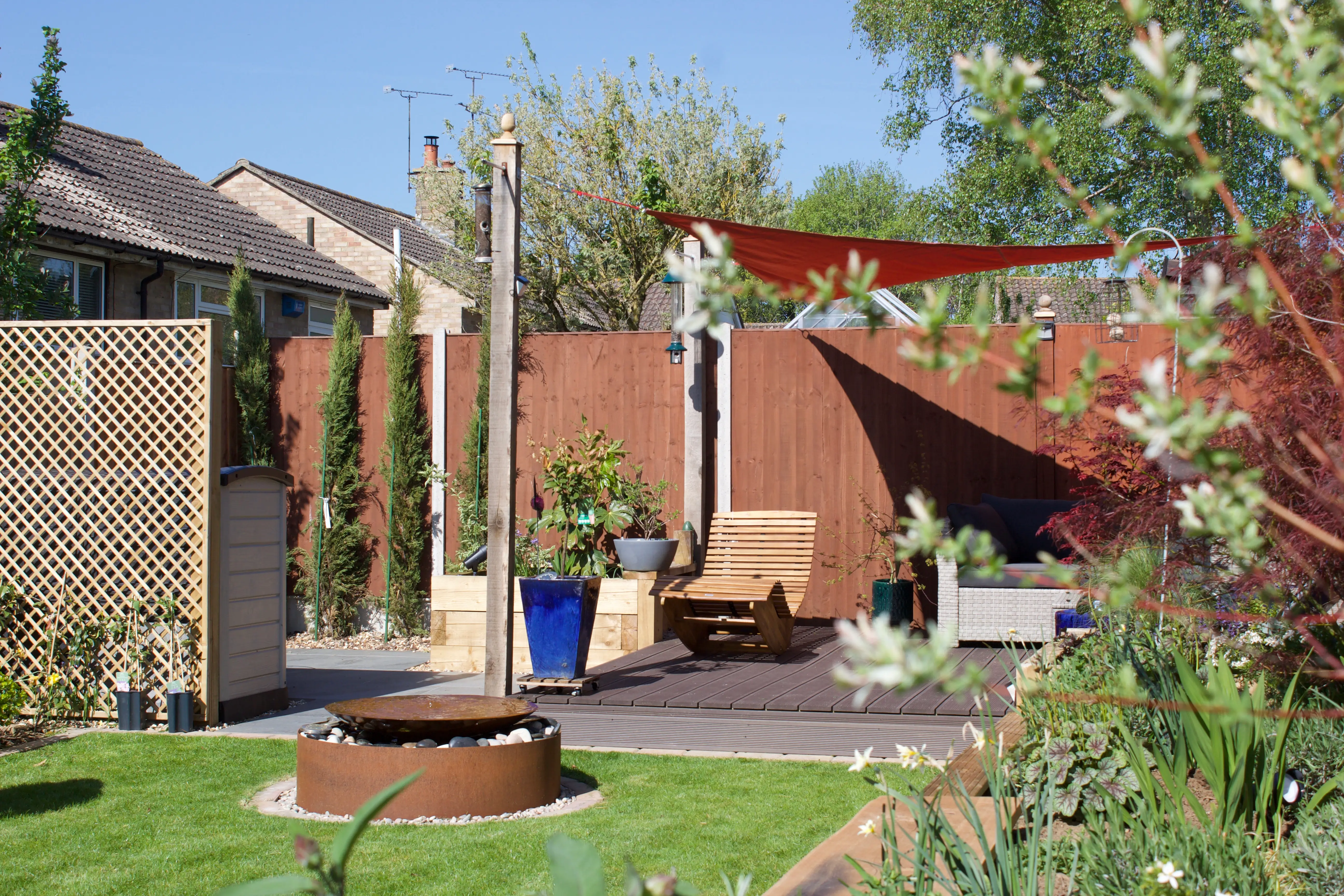 A sunny garden scene featuring landscaped areas, wooden fencing, potted plants, and modern outdoor furniture.