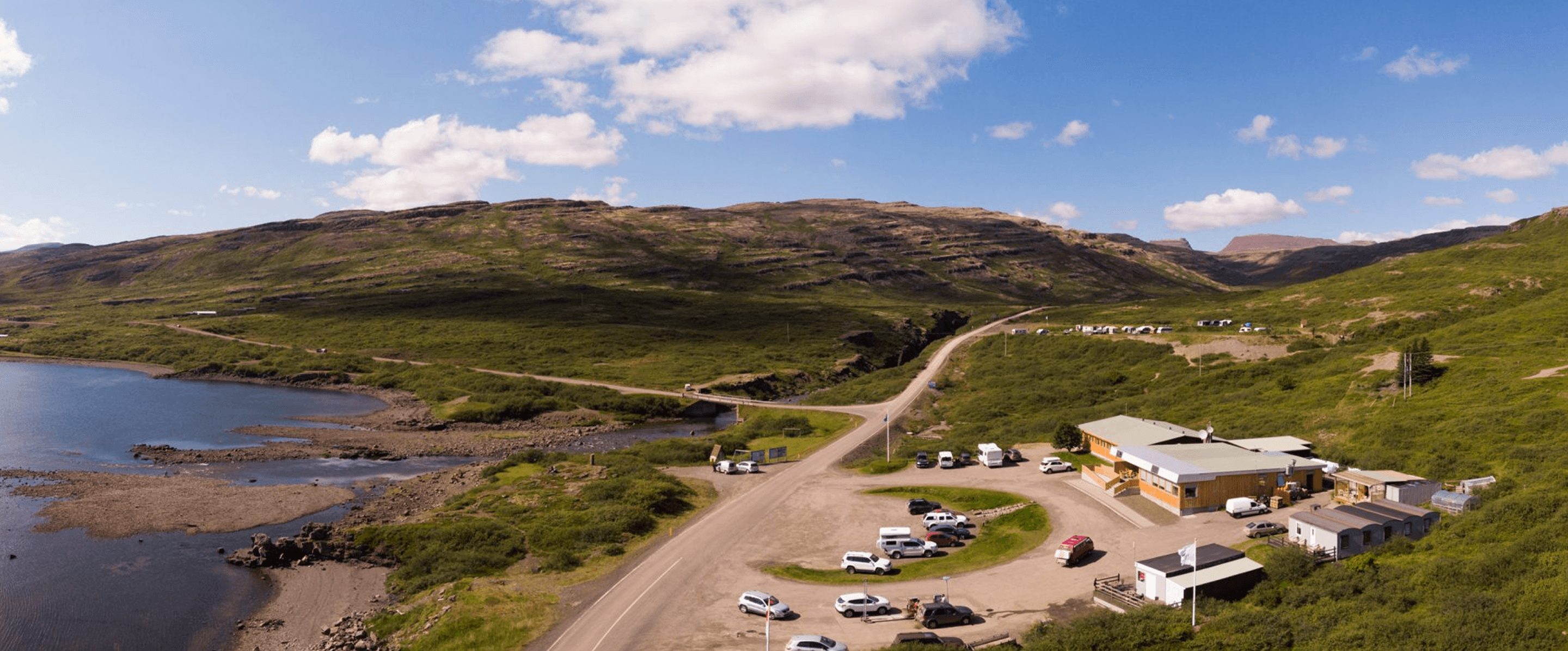 Winding road through green hills with parked cars and small buildings beside a river in a wide rural landscape.
