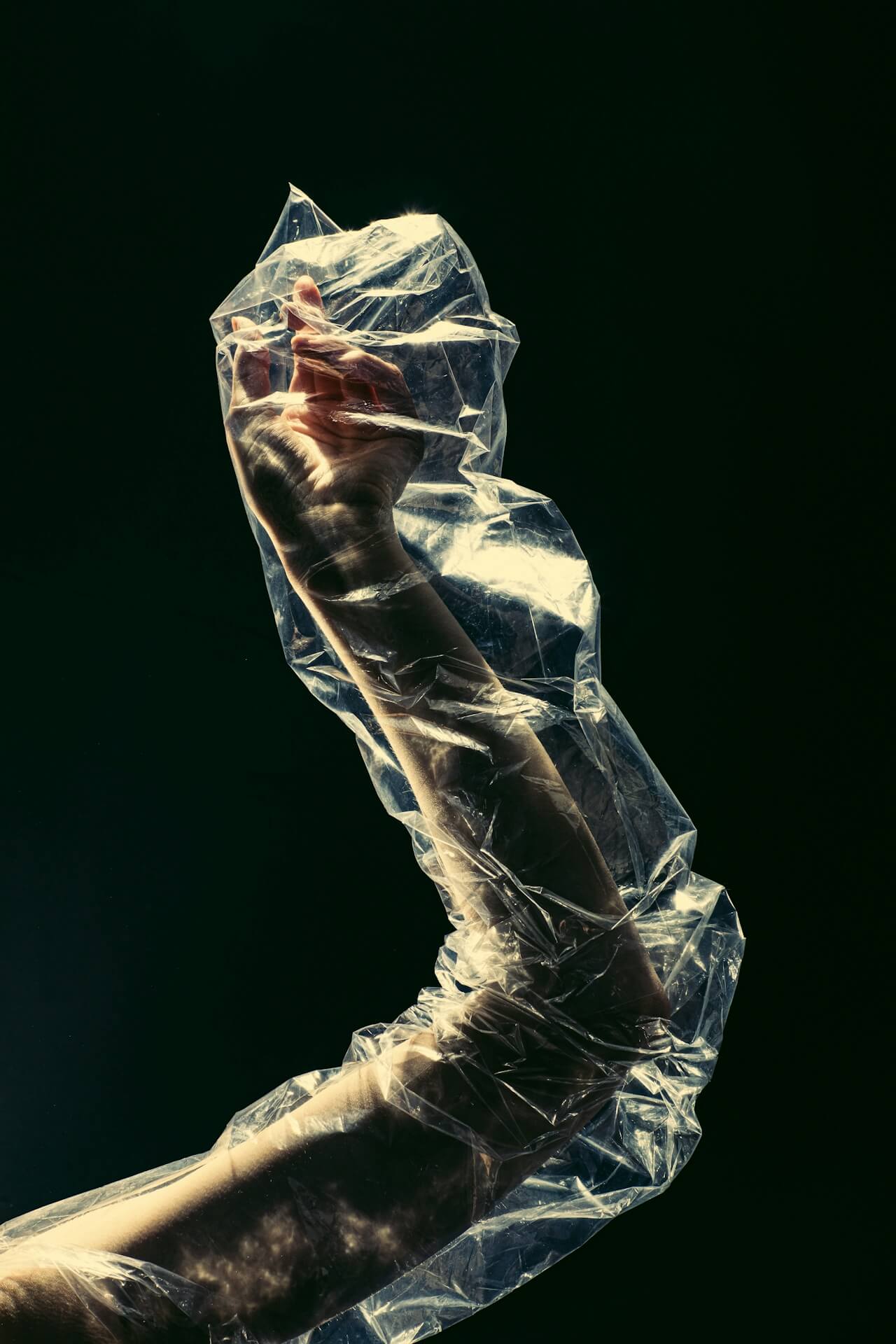 A hand wrapped in crinkled, translucent plastic reaches upwards against a dark background. The light highlights textures, evoking themes of entrapment and fragility.