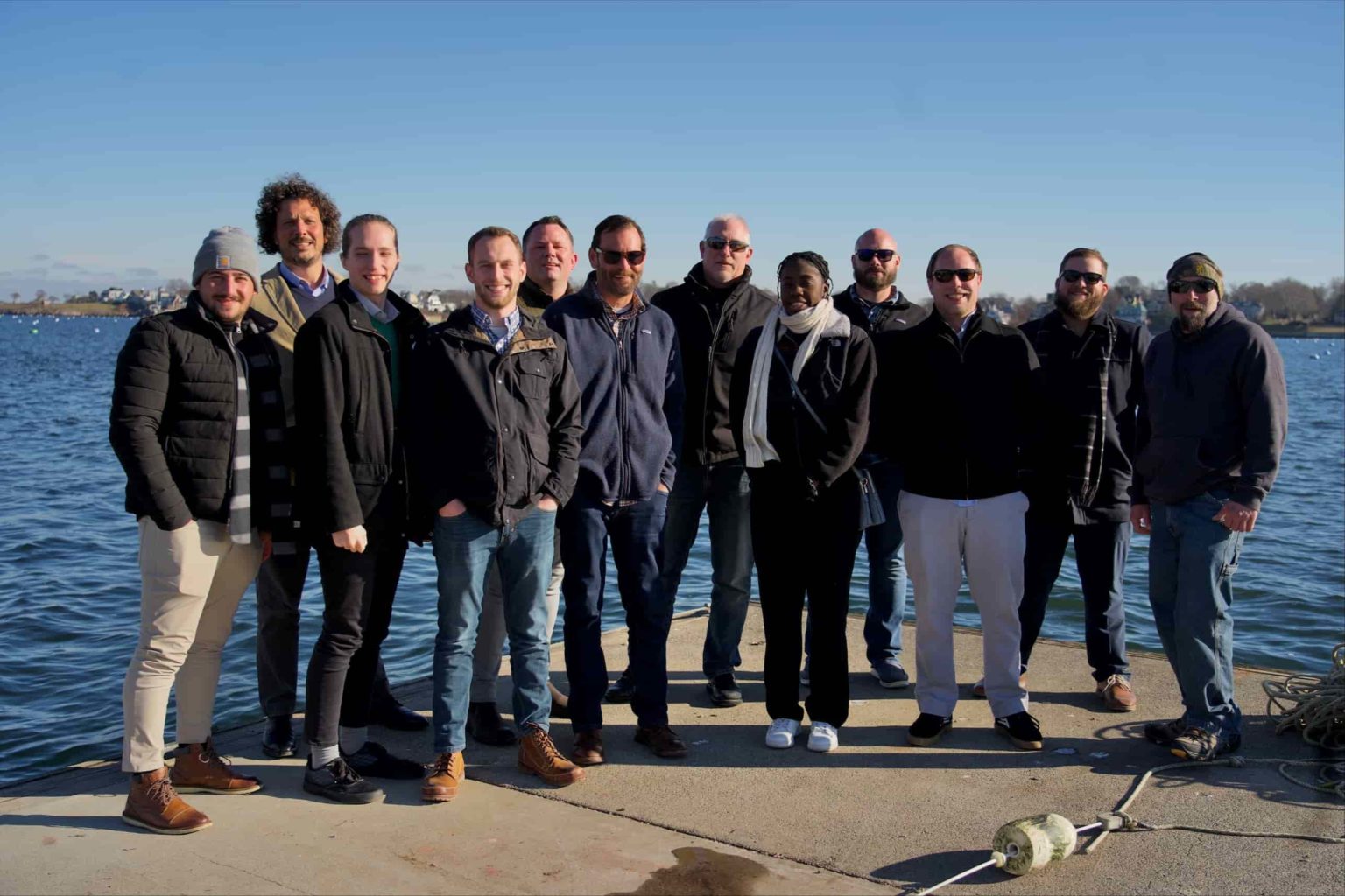 Group of eleven team members from NEDC New England Design and Construction standing on a waterfront pier on a sunny day. They are dressed casually in winter attire, with the blue harbor waters and distant shoreline visible behind them.