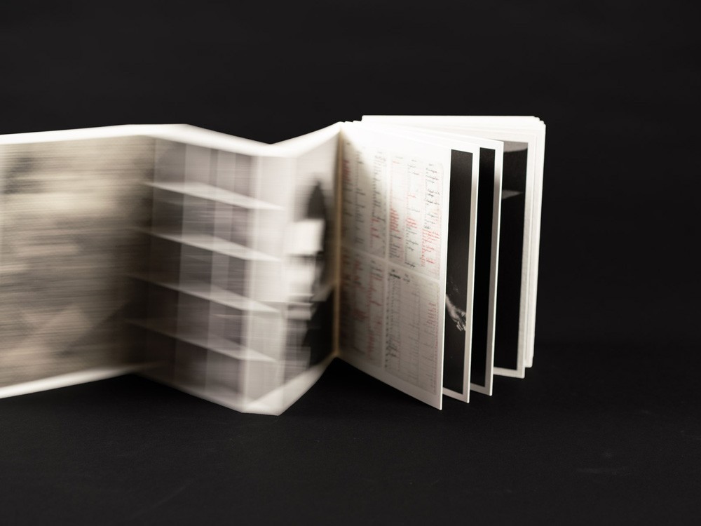 Een dynamische foto van een harmonica-gevouwen boek (leporello) op een zwarte ondergrond. De linkerzijde van het boek is vervaagd door beweging (motion blur), wat een suggestie van snelheid geeft, terwijl de rechterpagina's scherp zijn en lijsten met tekst en zwart-wit foto's tonen.