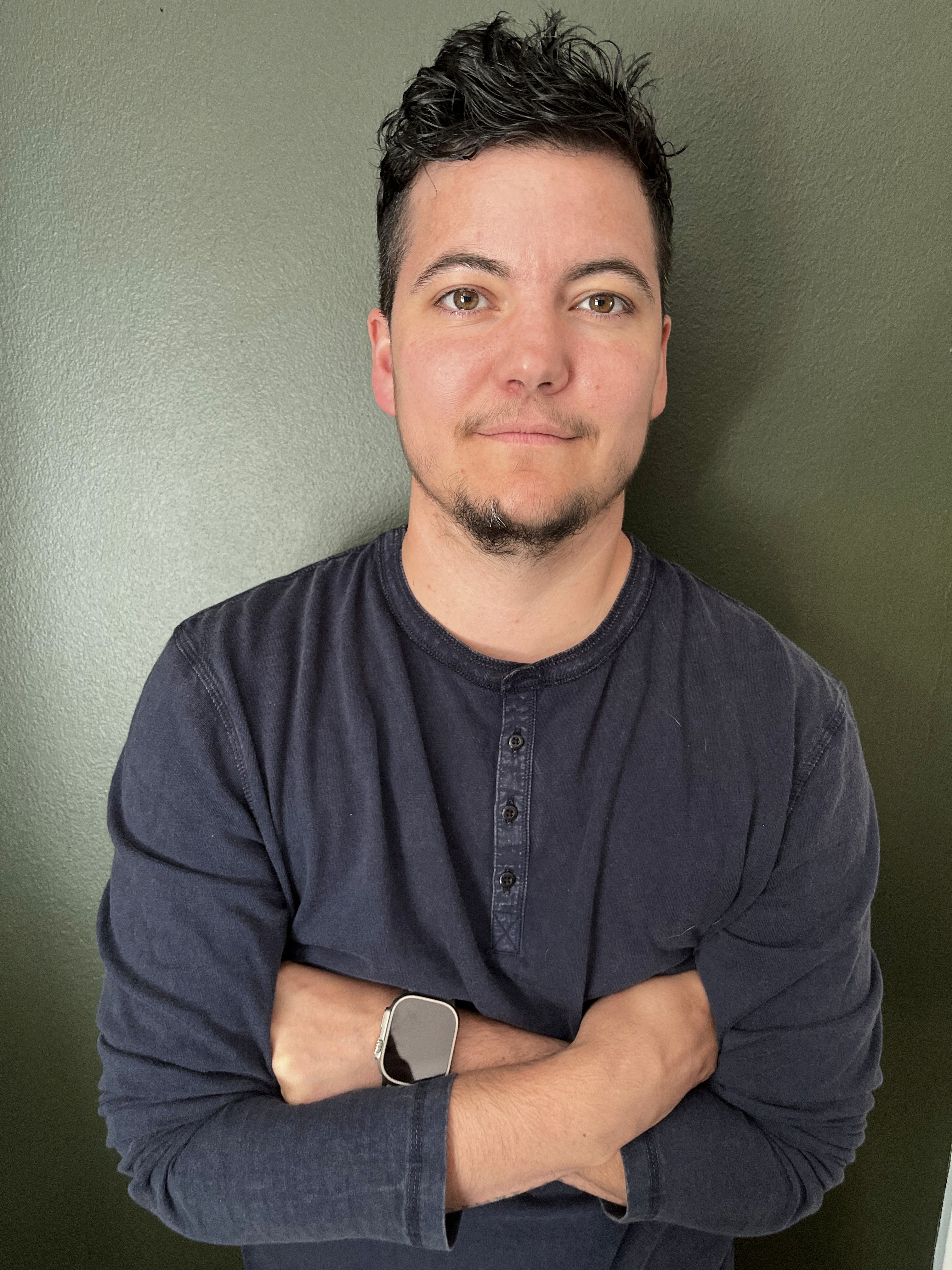 A person with short dark hair and a goatee stands with arms crossed, smiling against a green background.