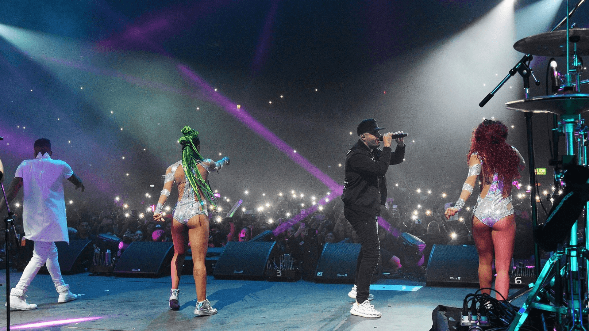 Cantante con gorra negra y micrófono actúa con tres bailarines brillantes; público levanta celulares iluminados bajo luces azules y púrpuras.