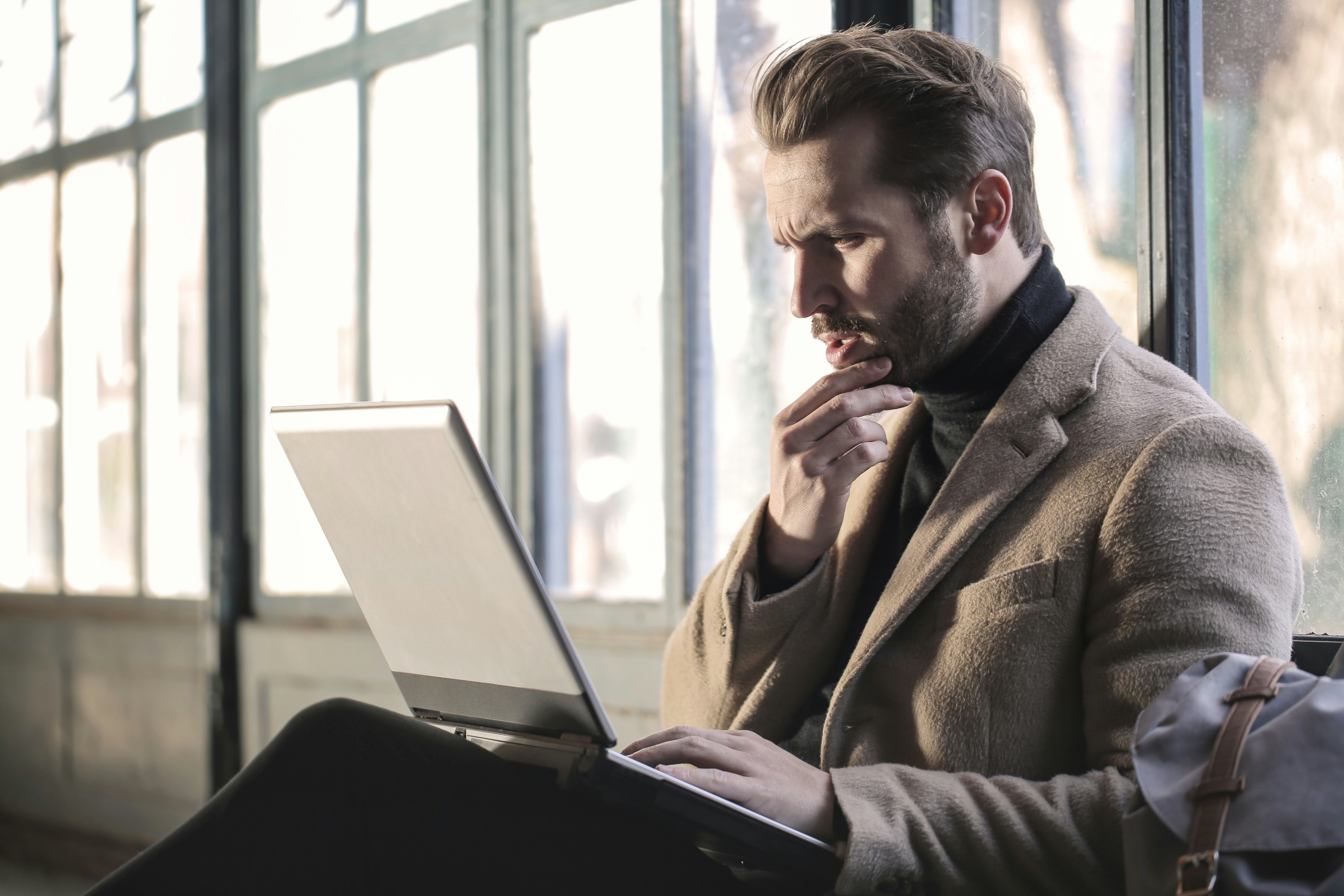 A well-dressed man is sat looking at his laptop, which sits on his knees.