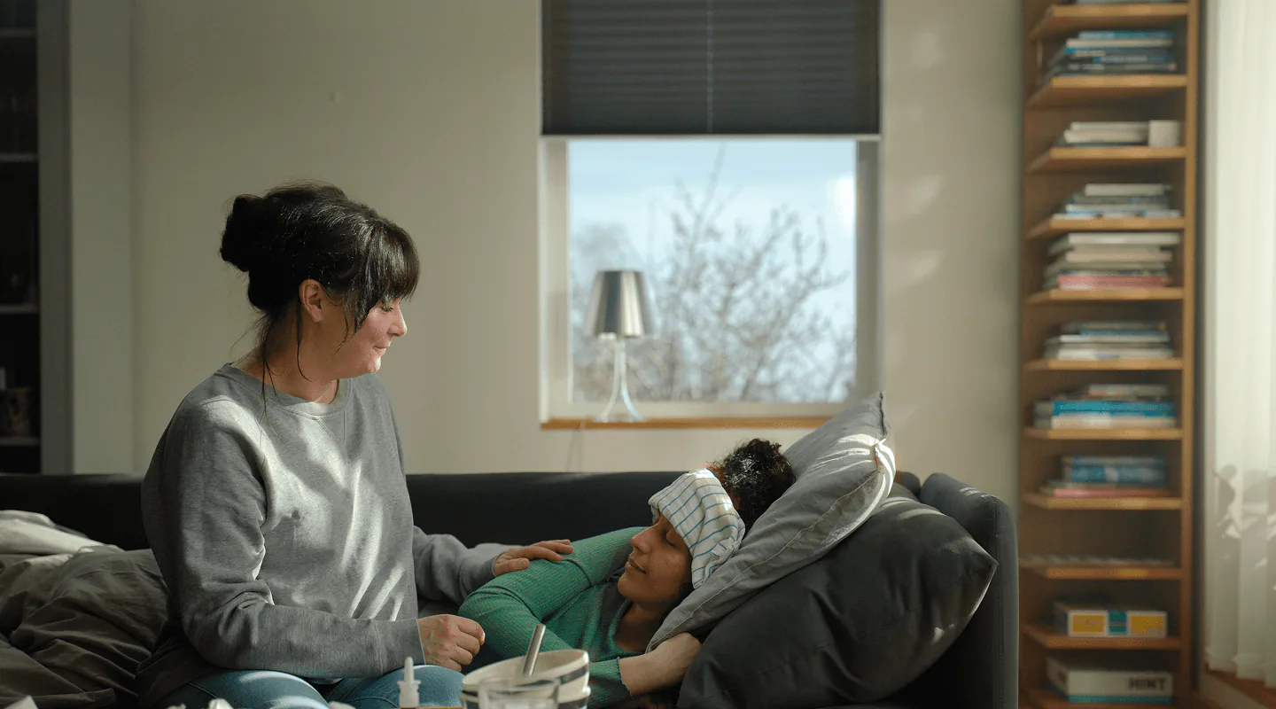 Kvinna sitter bredvid ett barn som ligger i en soffa med en mössa på, ur caset Där du är